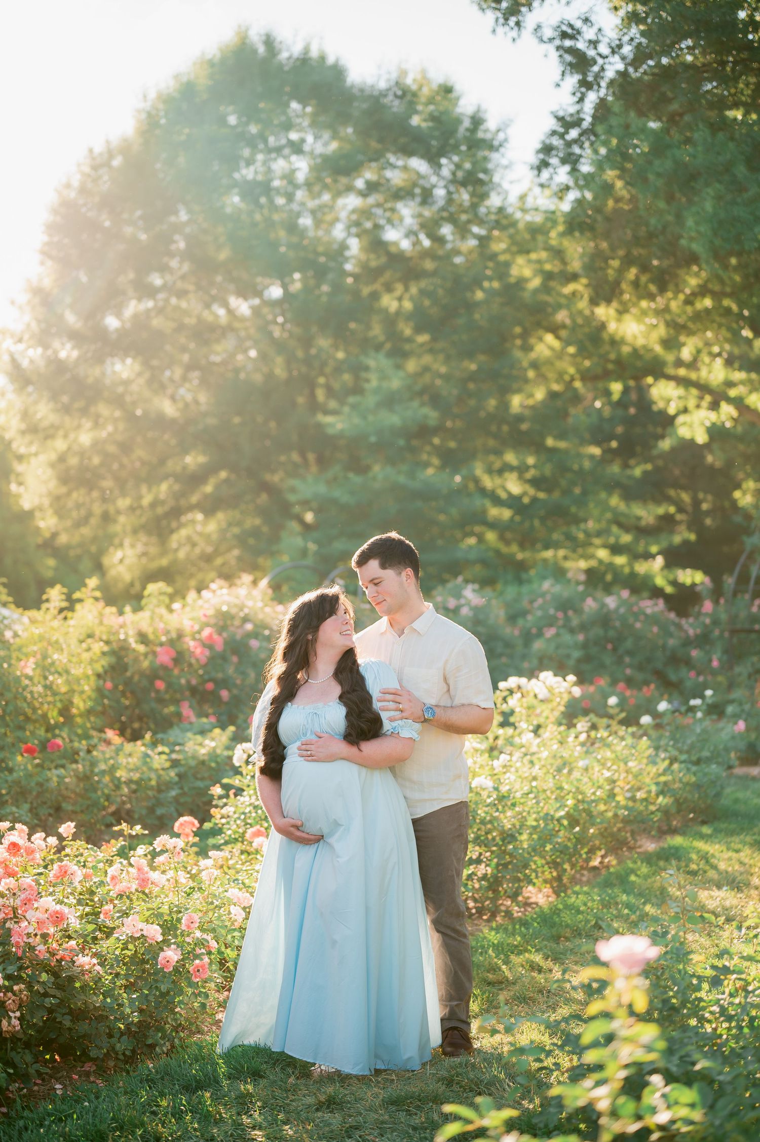 Pregnant couple embracing in a sunlit rose garden, woman in flowing blue gown, golden hour light filtering through trees.