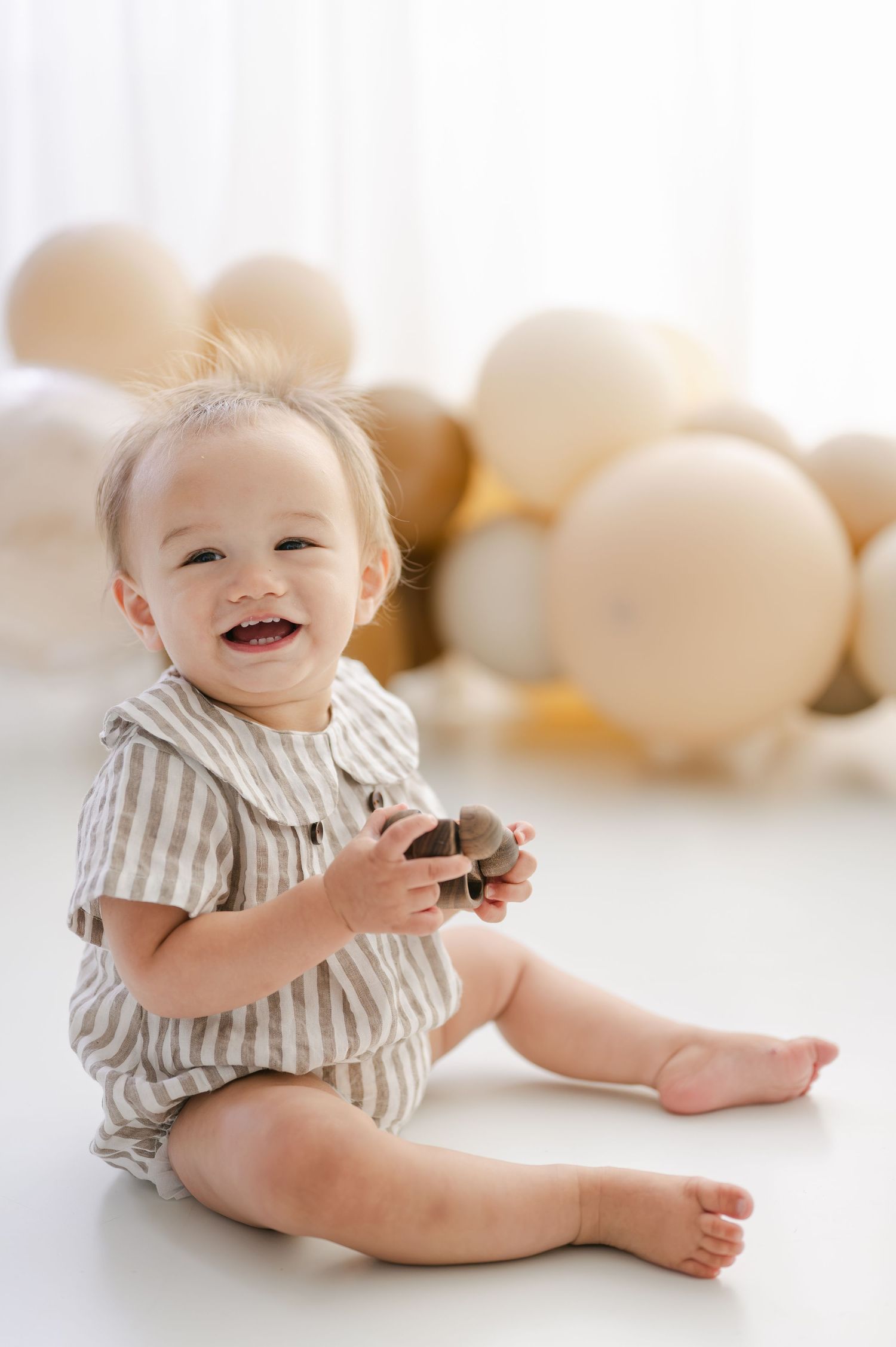 Smiling baby in striped romper sitting on white floor holding toy surrounded by beige and gold balloons.