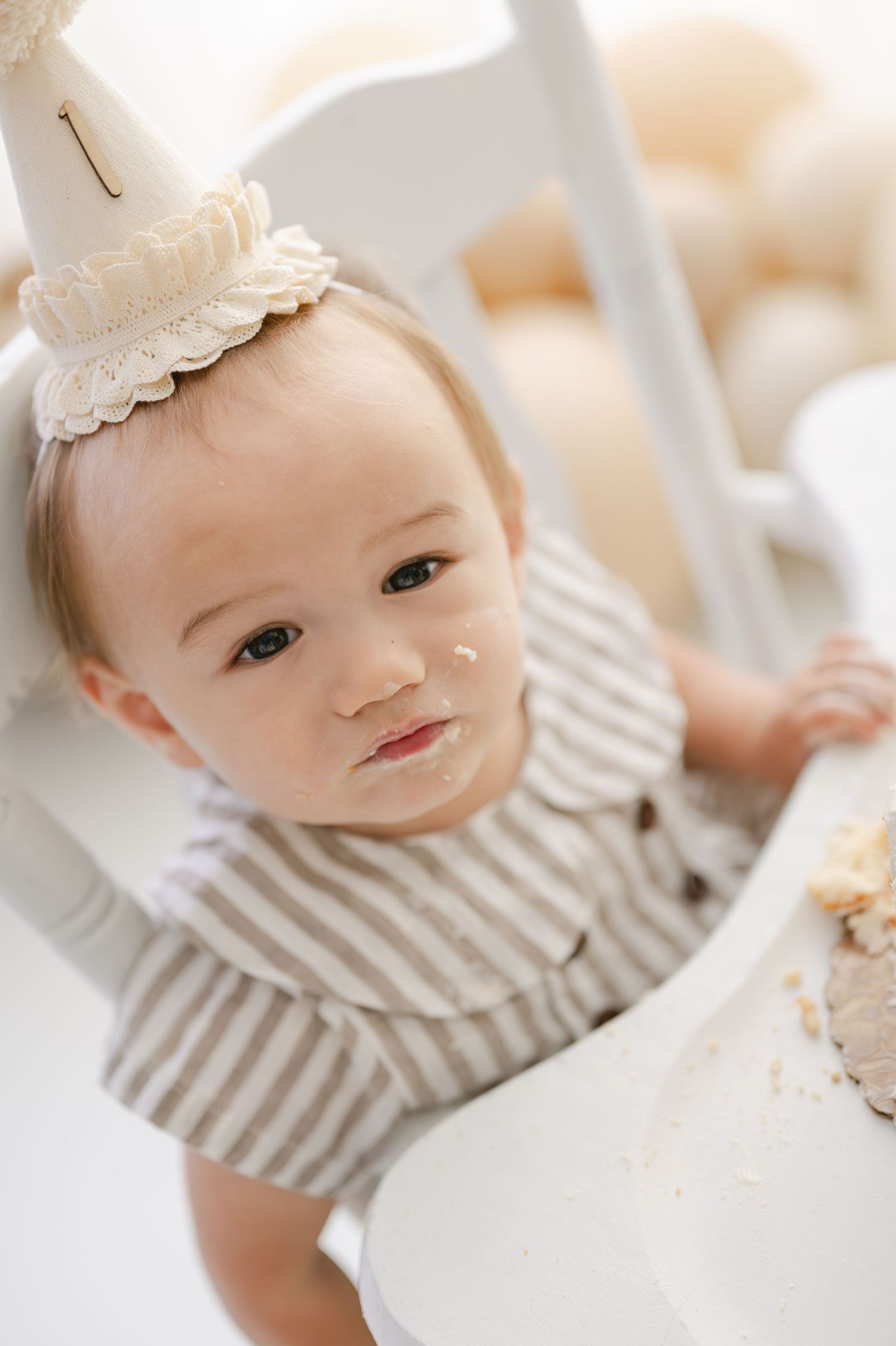 Cute baby in striped outfit and birthday hat sits in high chair with cake smash mess on face.