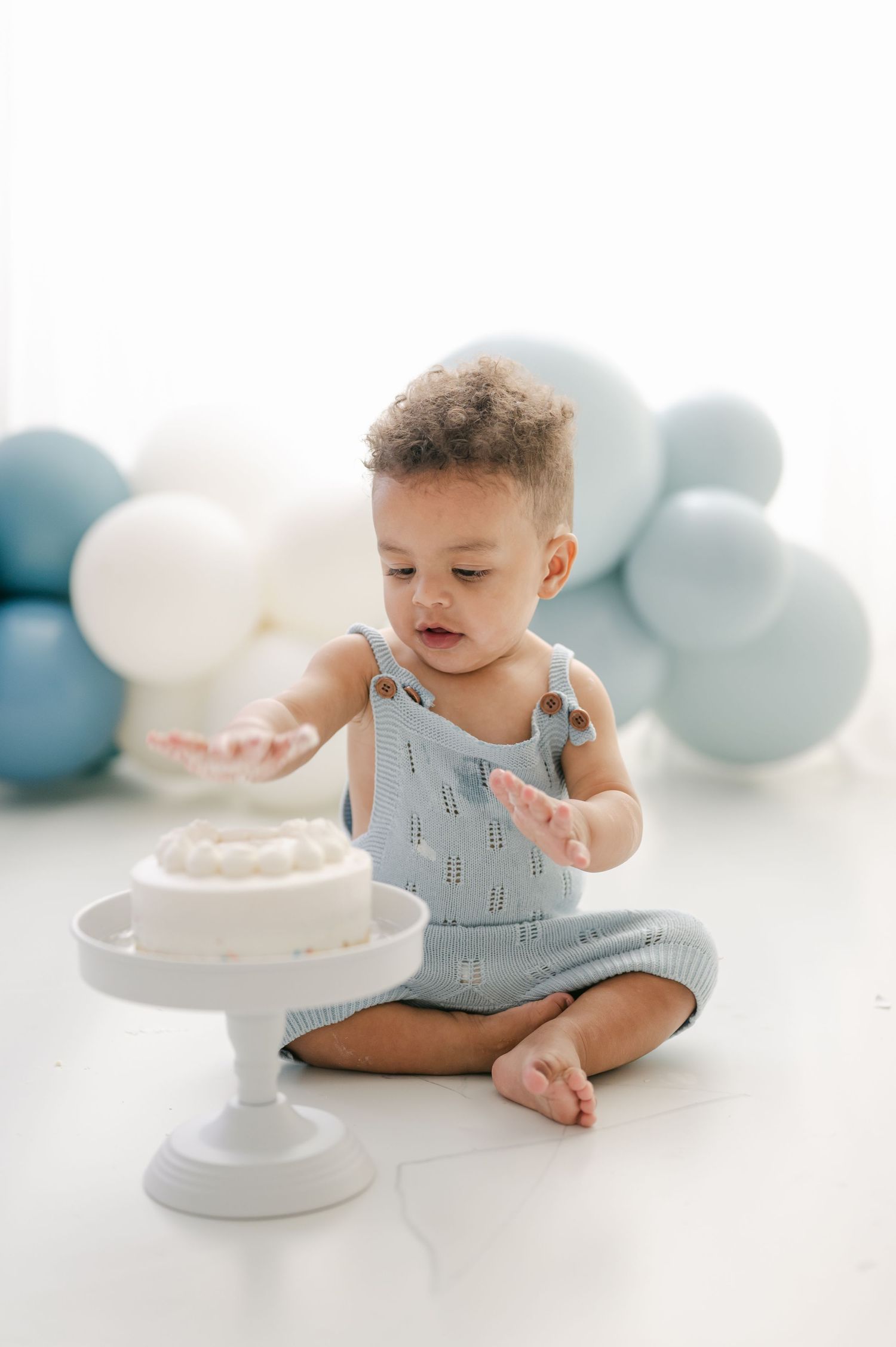 A baby boy in a blue knit romper sits on a white floor reaching toward a smash cake, with blue and white balloons behind him.