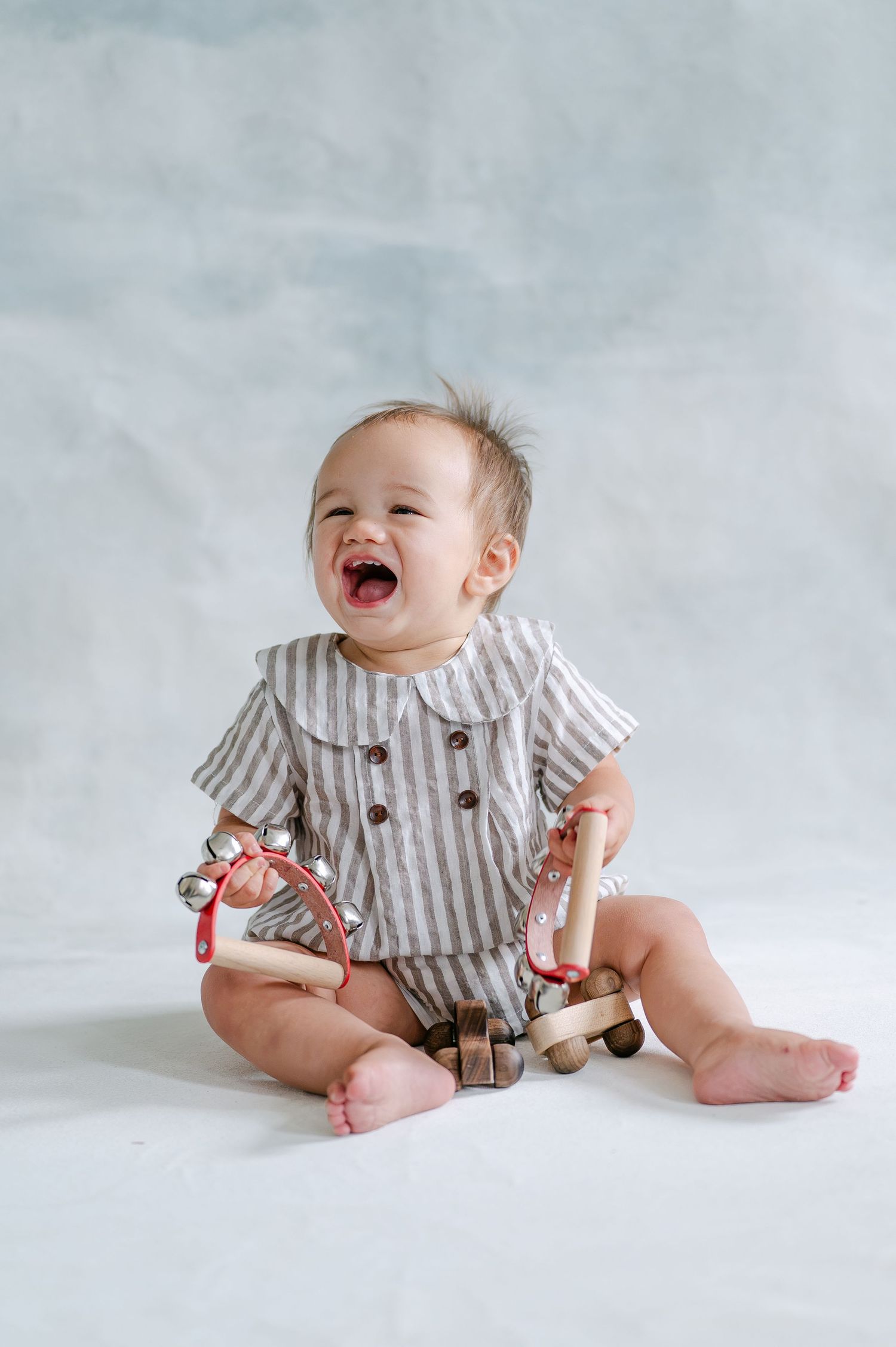 Laughing baby in striped romper sitting on white floor holding wooden toys and tambourine.