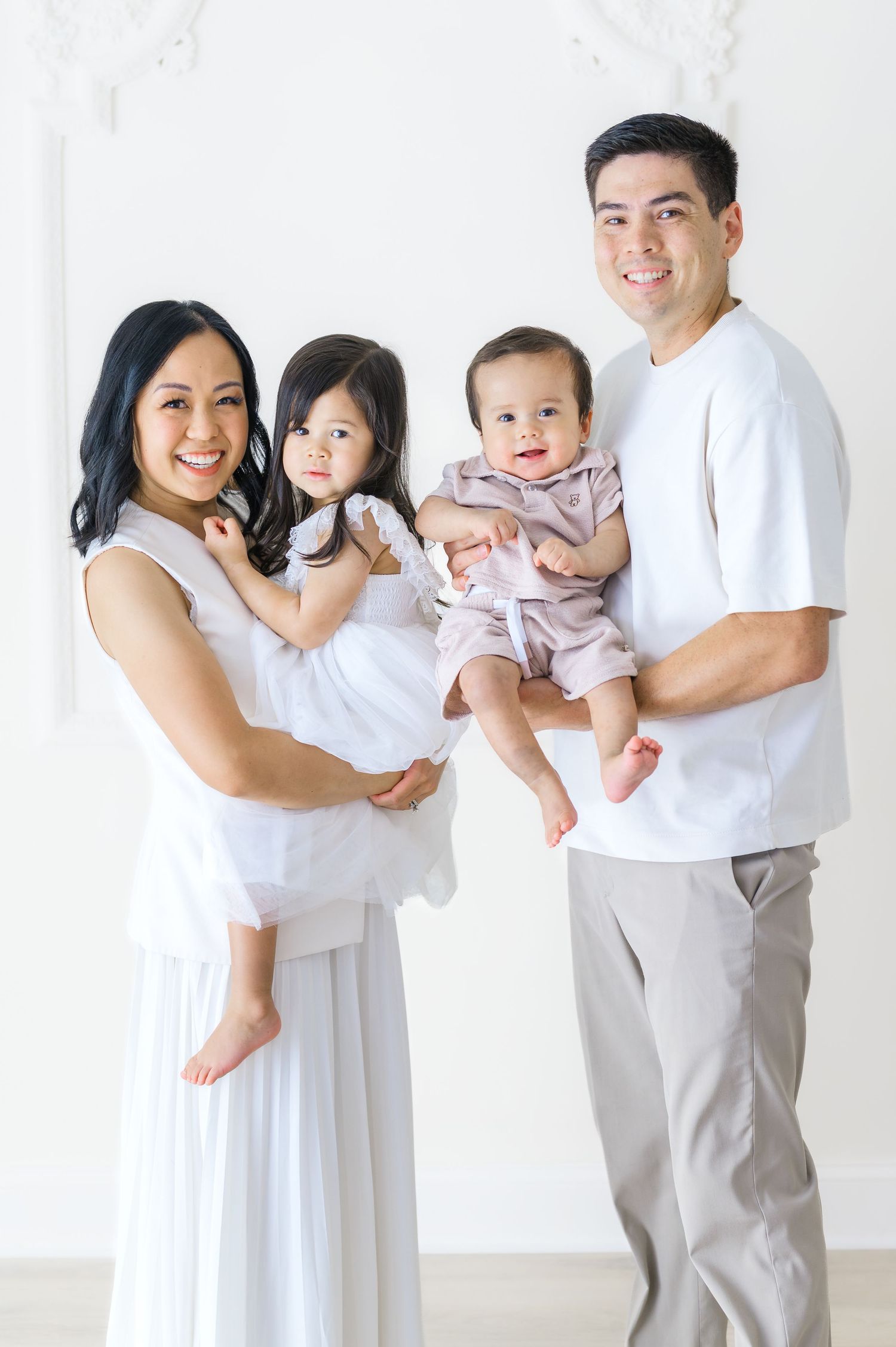 A happy Asian family of four poses in a bright white studio, parents holding two young children dressed in soft pastel outfits.