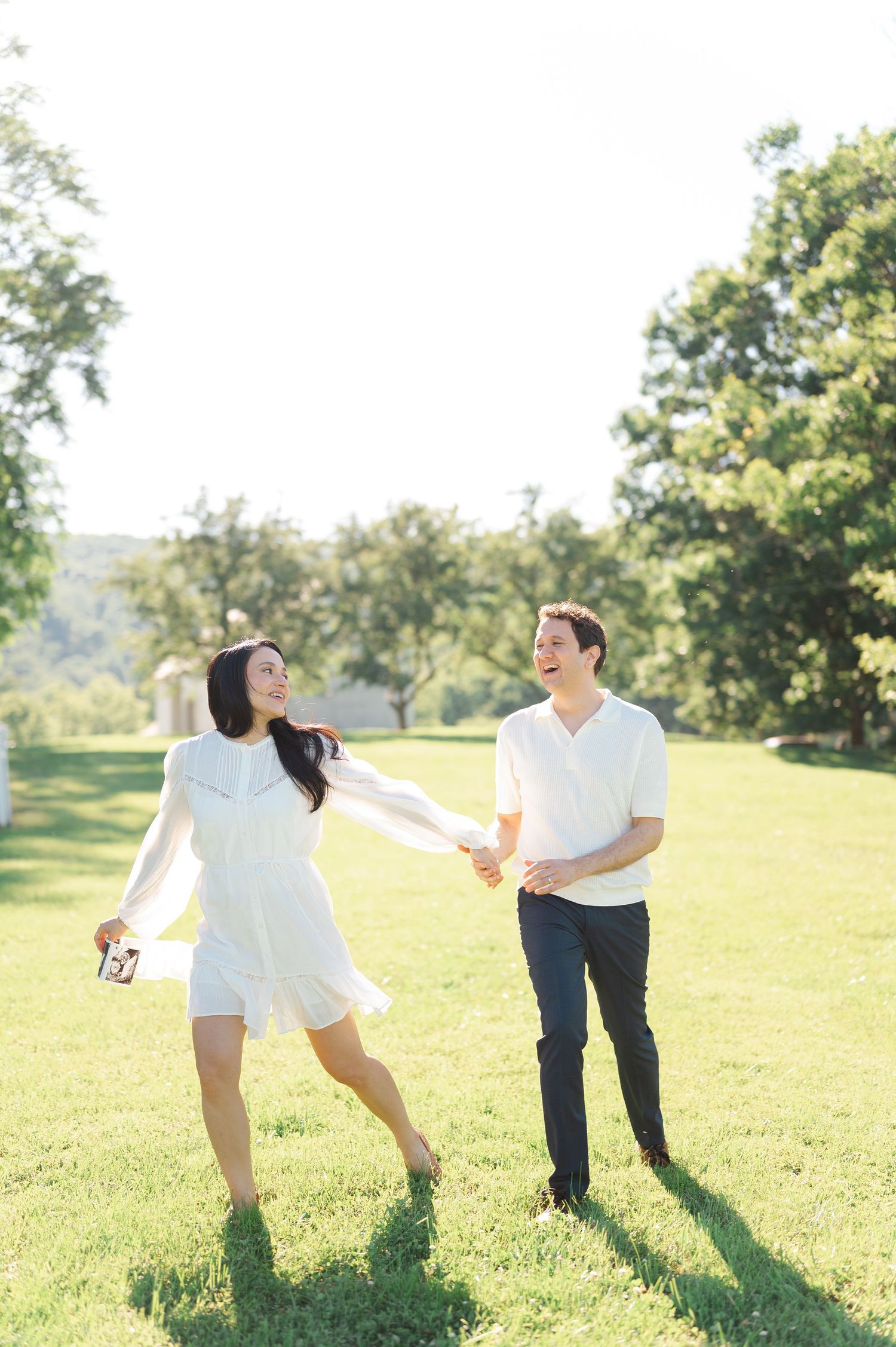 A joyful couple holds hands and dances in a sunny green park, the woman in a white dress holding an ultrasound photo.