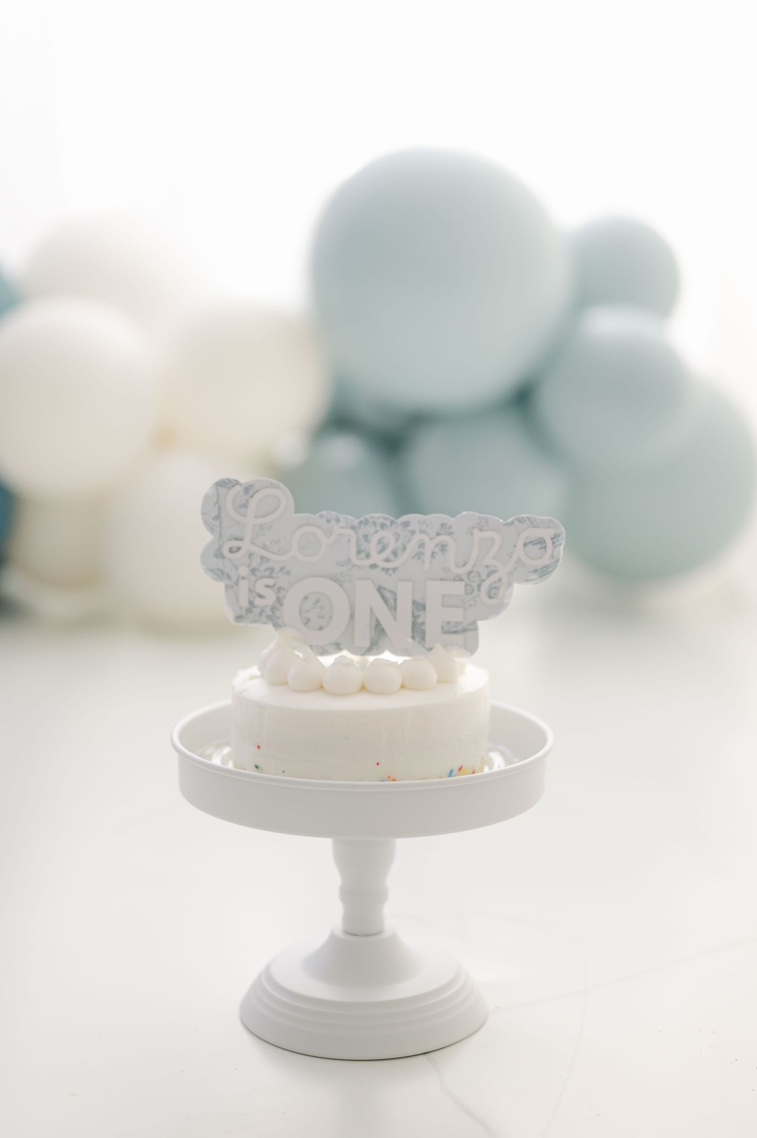 White smash cake with ONE topper on pedestal stand, soft blue and white balloons in background, first birthday celebration.