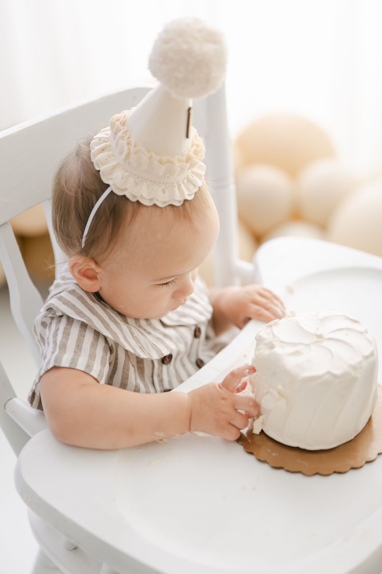 Baby in birthday hat smashes first cake in white high chair, surrounded by neutral balloons.
