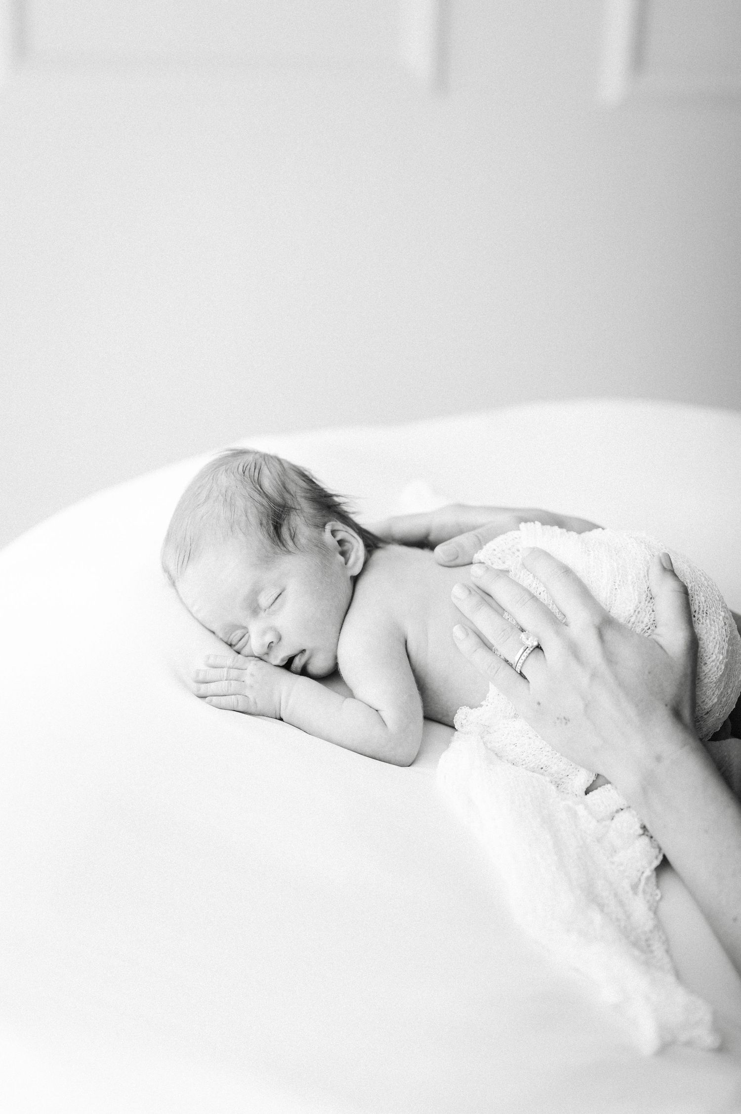 Black and white photo of a sleeping newborn baby cradled gently by a parent's hand with a wedding ring.