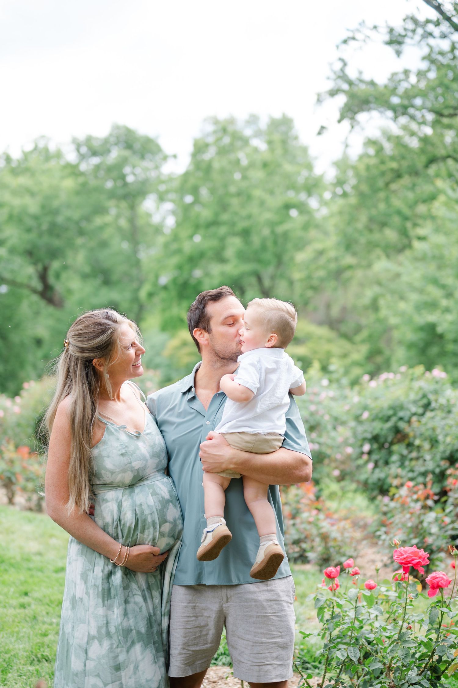 Pregnant woman in sage dress stands with man kissing toddler boy in rose garden.