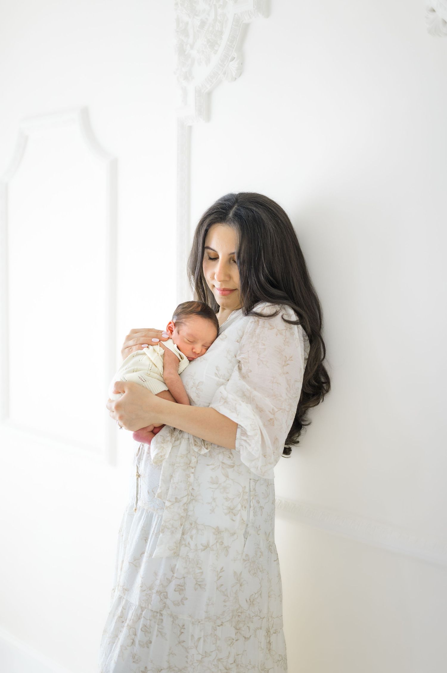 A mother in a white floral dress tenderly holds her sleeping newborn baby against her chest in a bright white room.