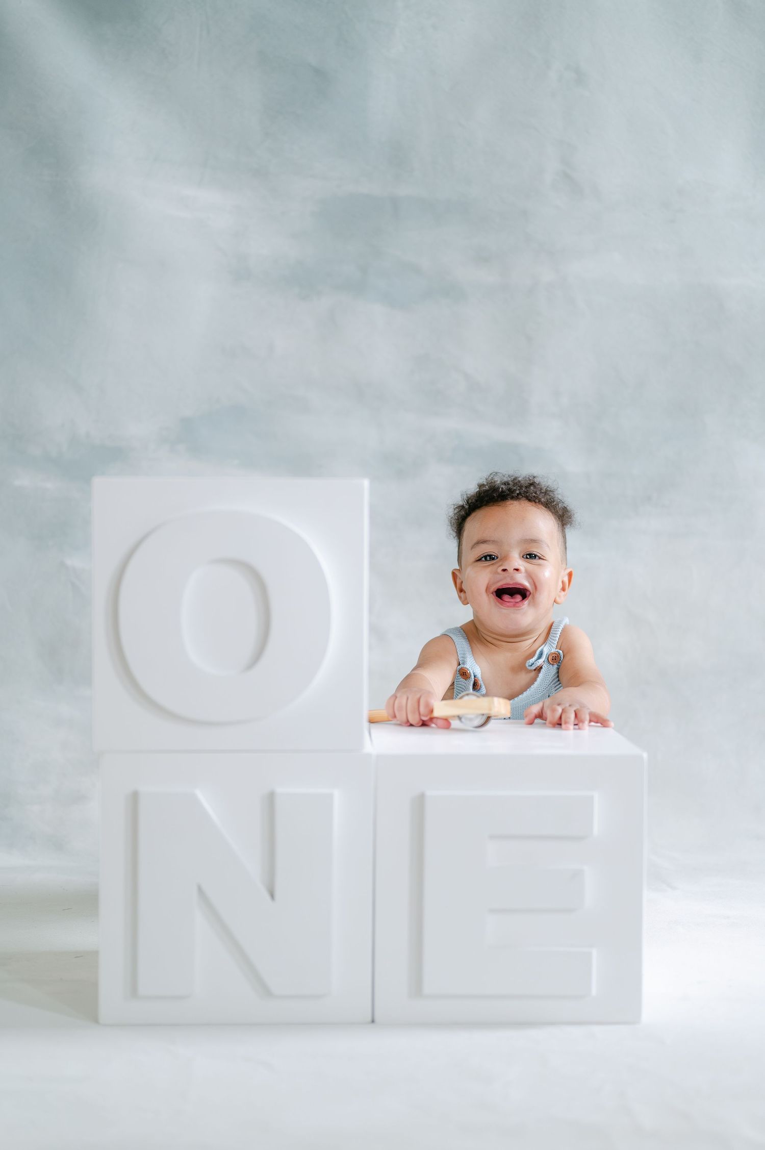 Smiling baby leaning on white letter blocks spelling ONE, celebrating first birthday against a soft gray background.