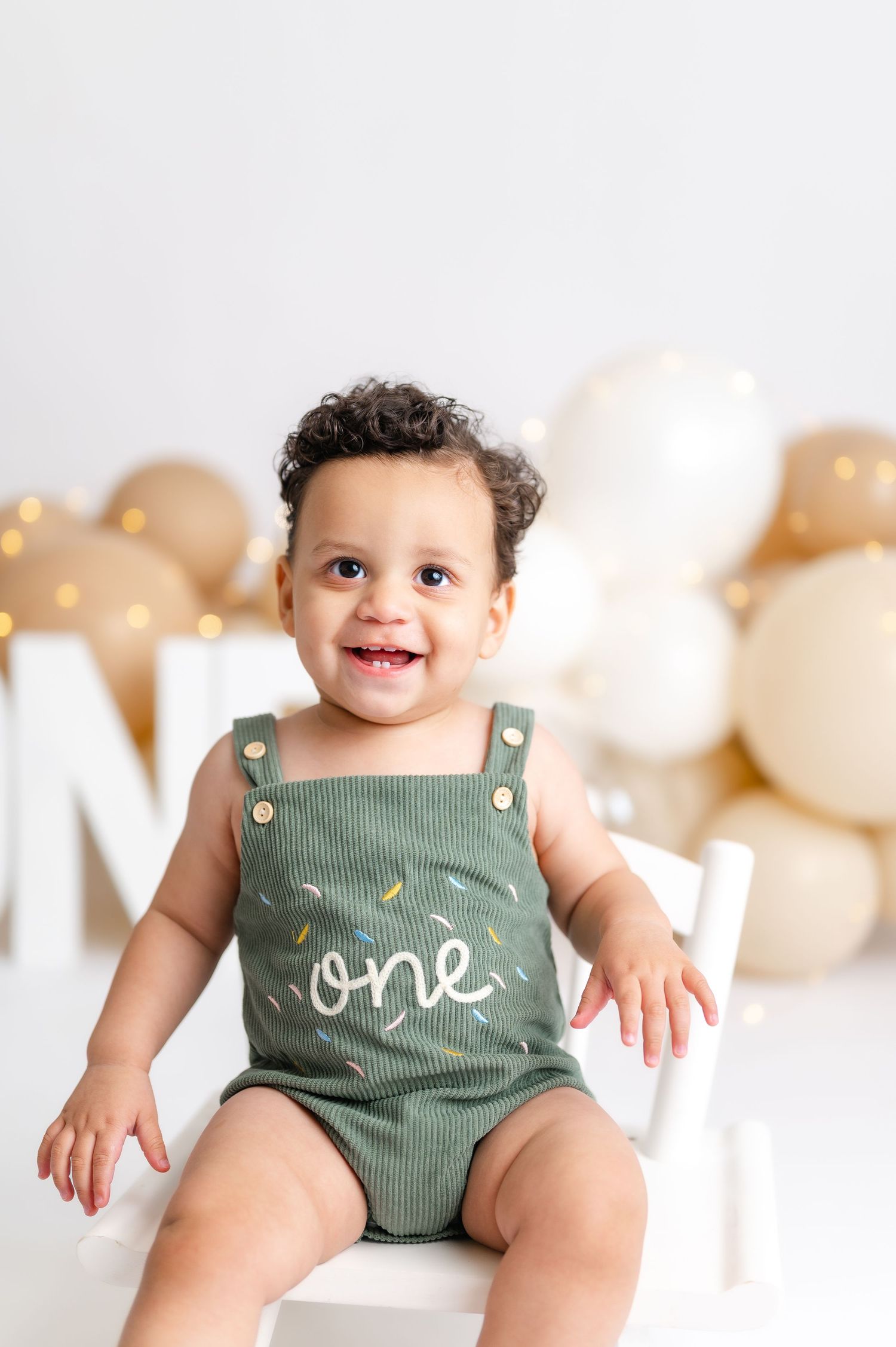 Smiling baby in green “one” romper sits on white chair with balloons for first birthday photo.