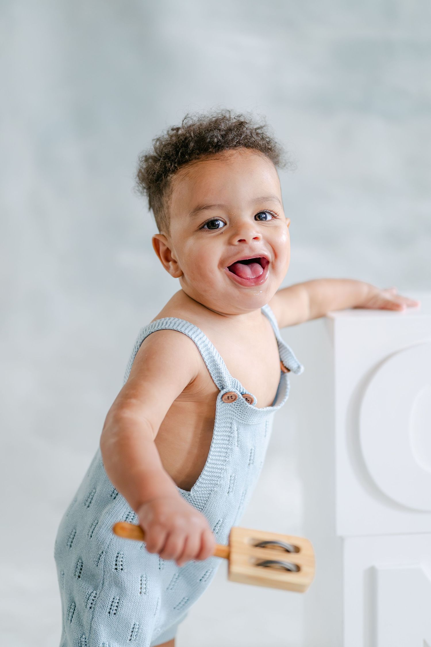 Happy baby in blue knit romper standing and holding a wooden toy, smiling joyfully in a bright studio.