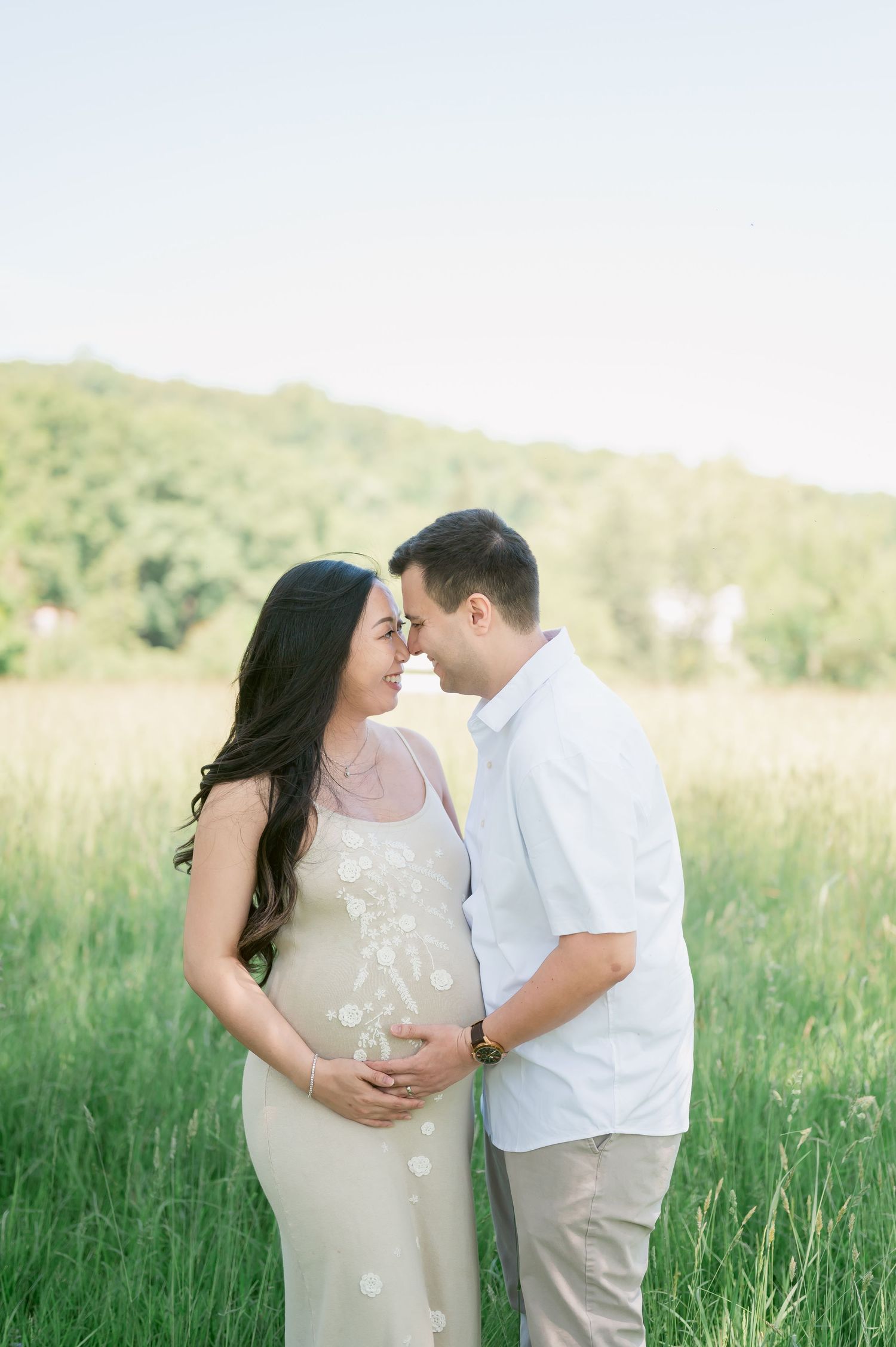 Couple sharing a tender moment in a sunlit field, pregnant woman in floral dress with partner in white shirt.