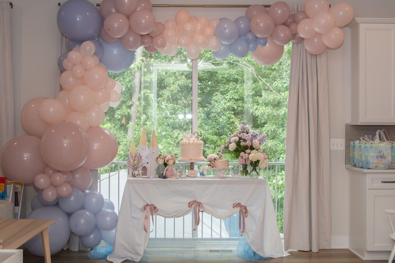 Pastel pink and purple balloon decorations adorn a white room with large windows and natural lighting.