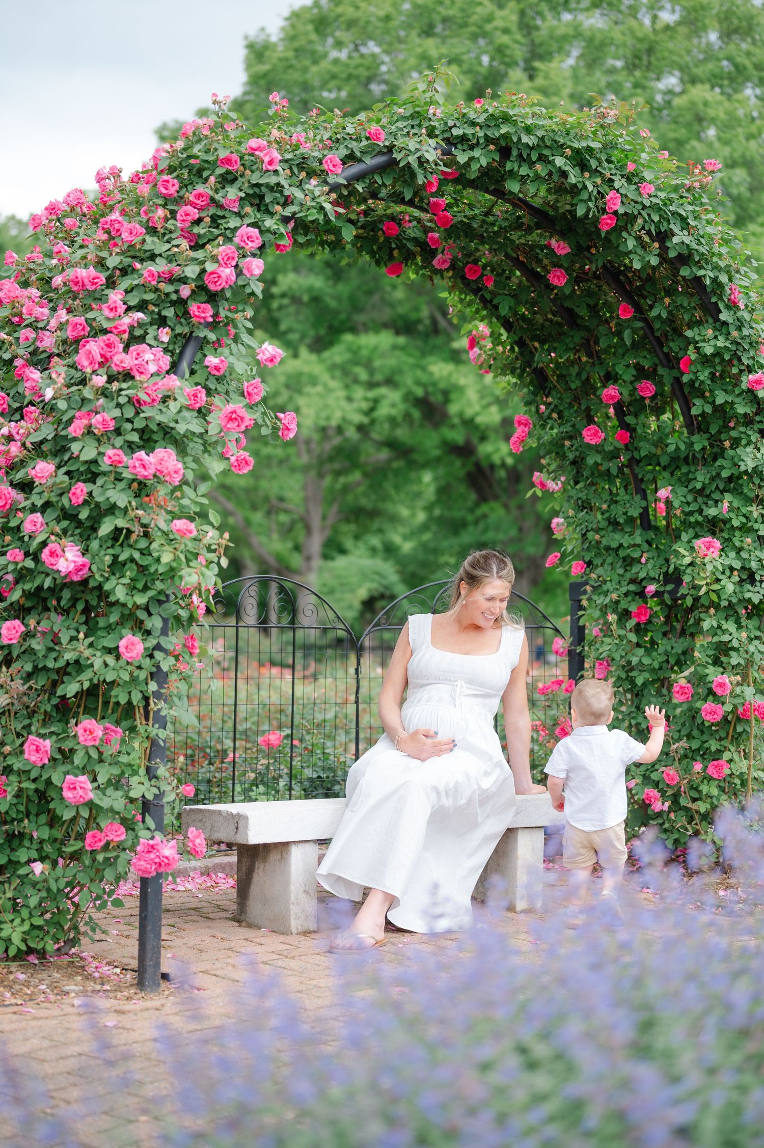 Pregnant woman in white dress sits on bench under pink rose arch with toddler in lush garden.