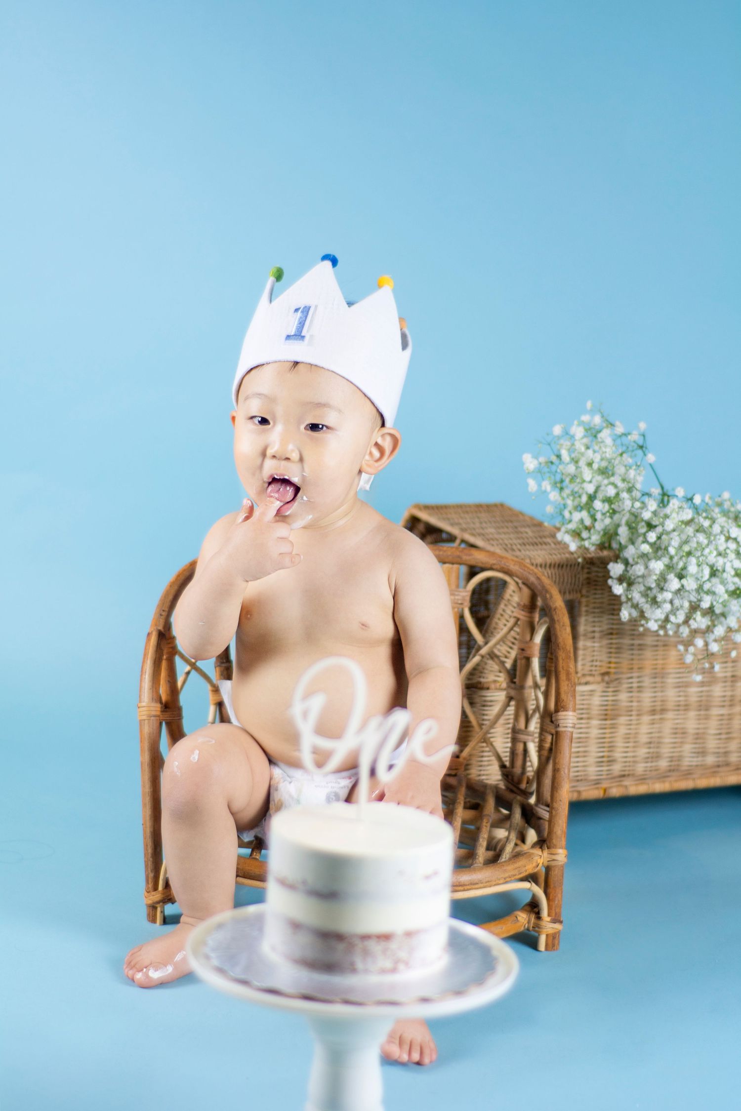 Baby wearing a blue crown enjoying their first birthday cake smash against a light blue backdrop.