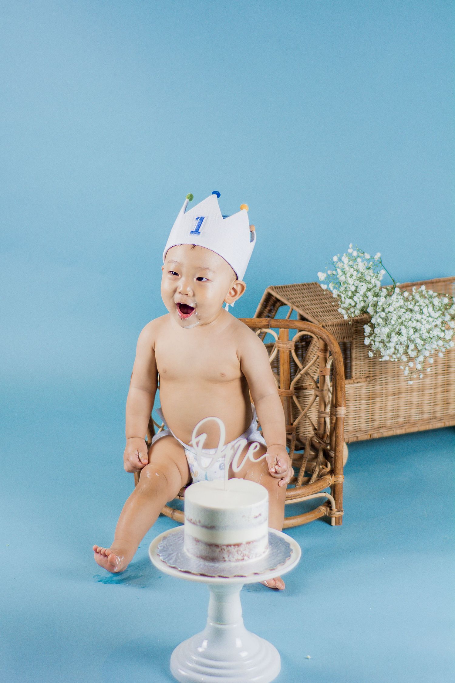 Baby in white paper crown excitedly approaches birthday cake on white pedestal against blue background.