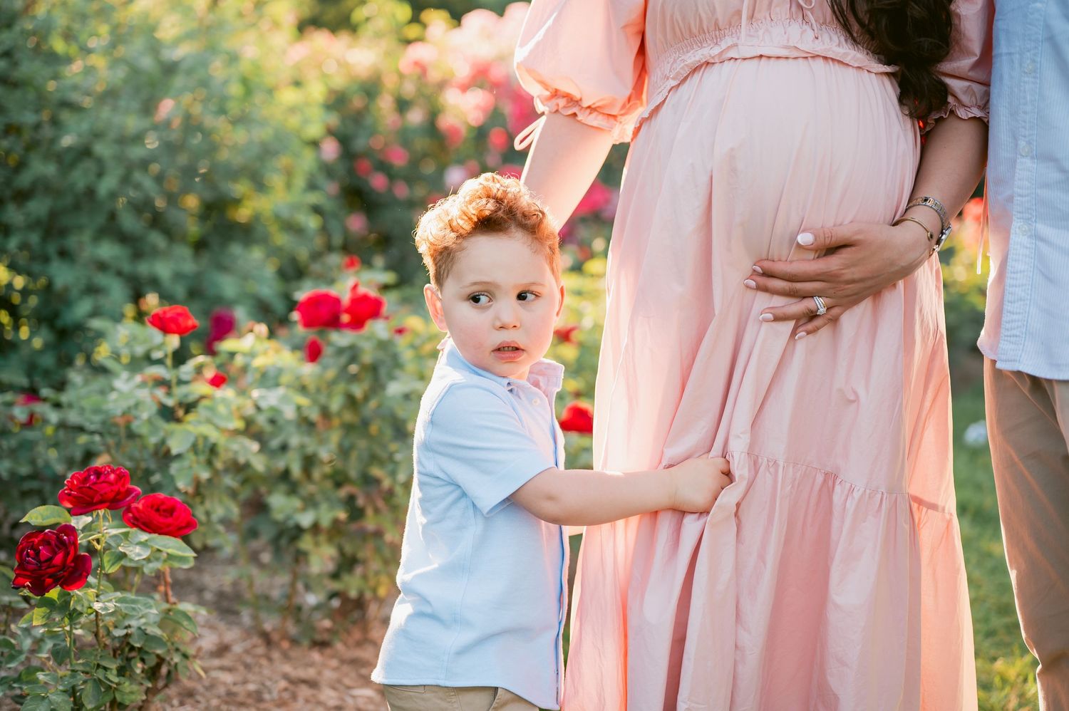 A young red-haired boy stands beside a pregnant woman in a pink dress among blooming red roses.