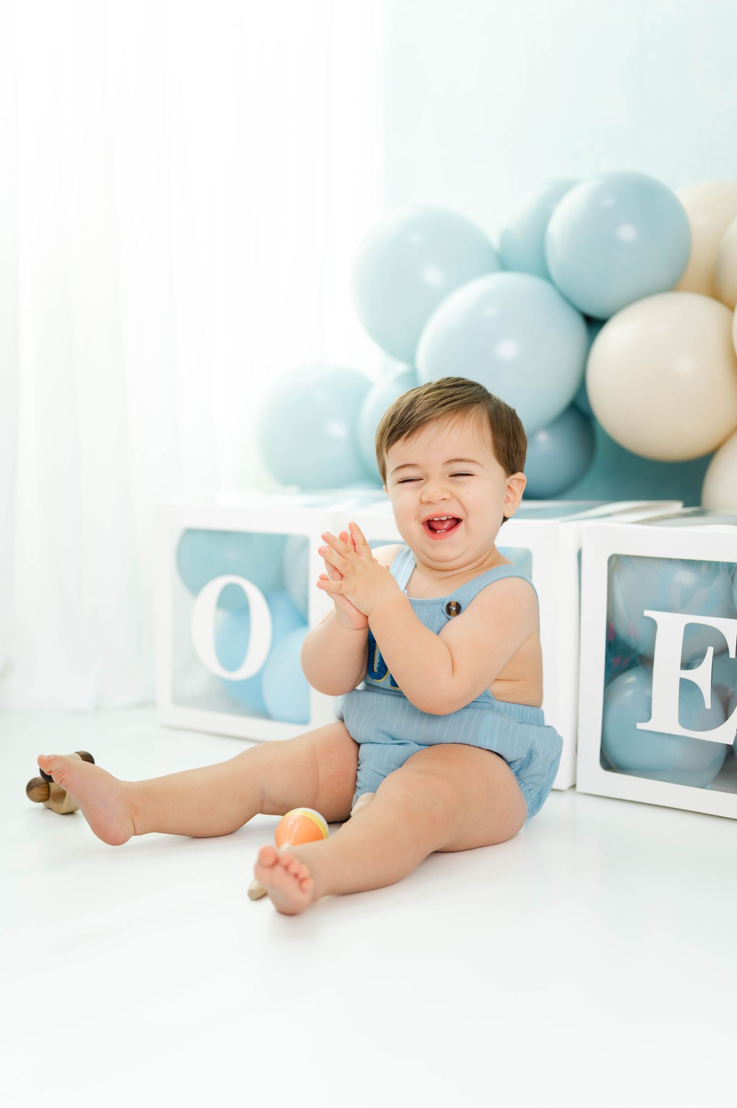 Baby in white diaper laughs while sitting on floor with blue and white balloons and letter decorations in background.
