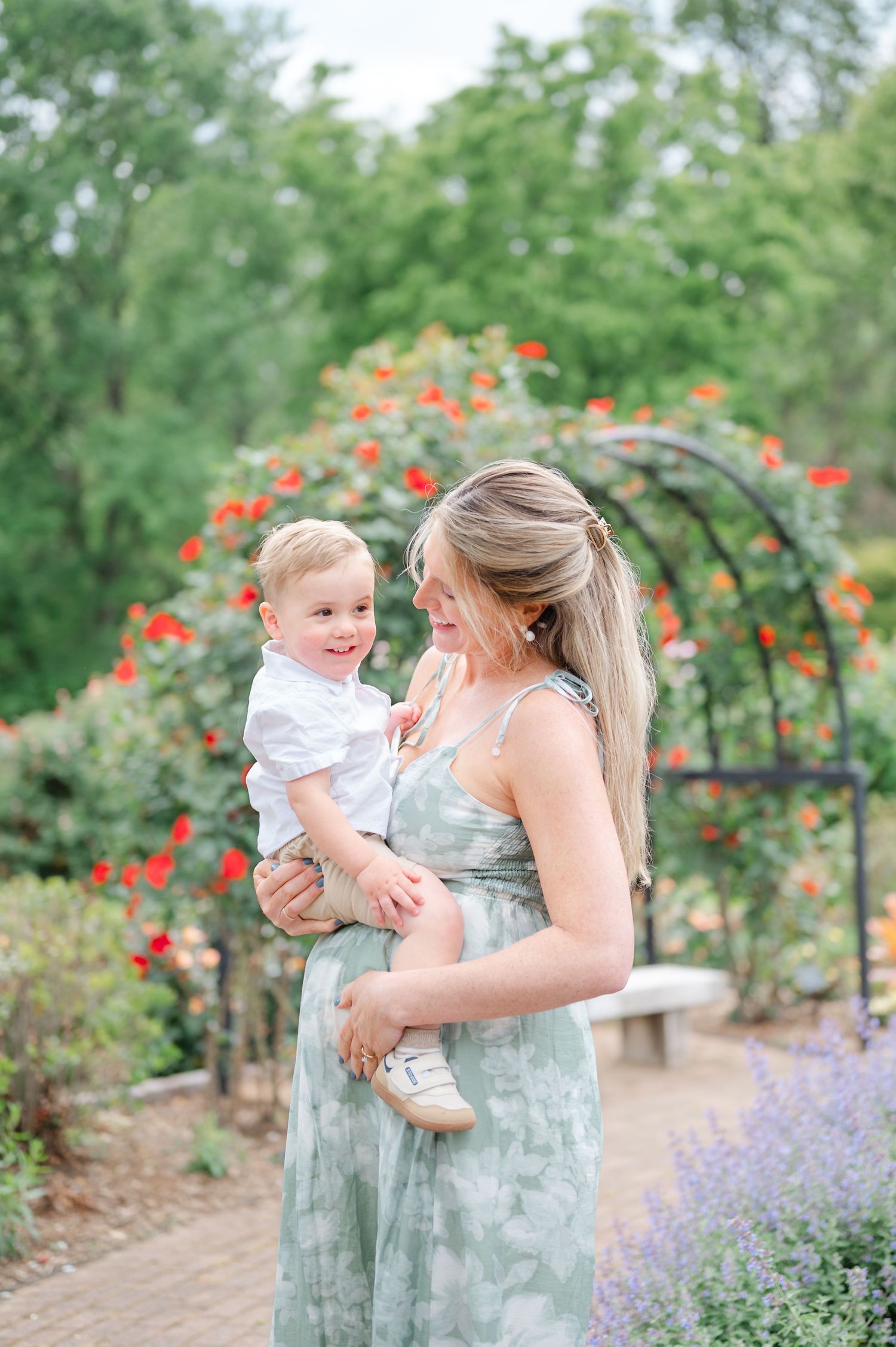 A mother in a floral dress holds her smiling toddler boy in a garden with red roses and an arch.