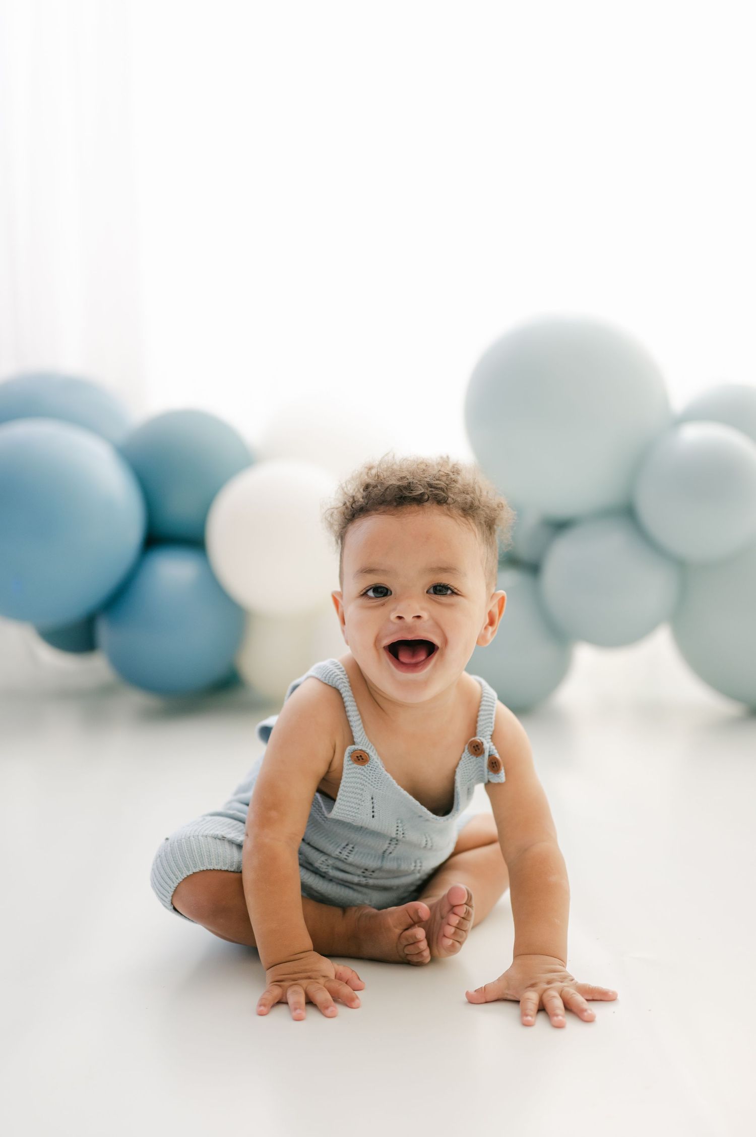 Smiling baby boy in blue romper sits on white floor surrounded by blue and white balloons.