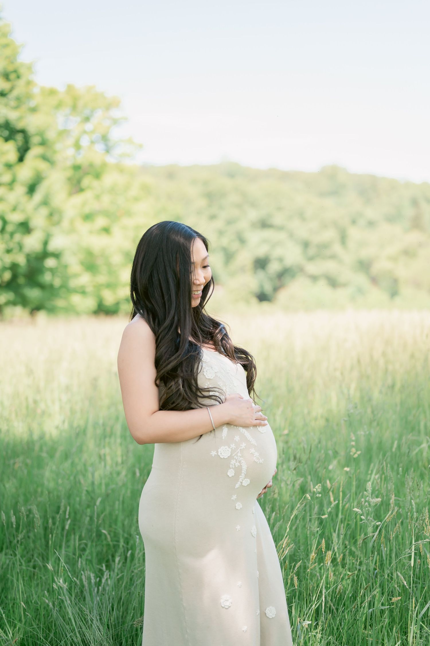 Pregnant woman in white floral gown standing in green meadow, cradling baby bump, surrounded by lush trees.