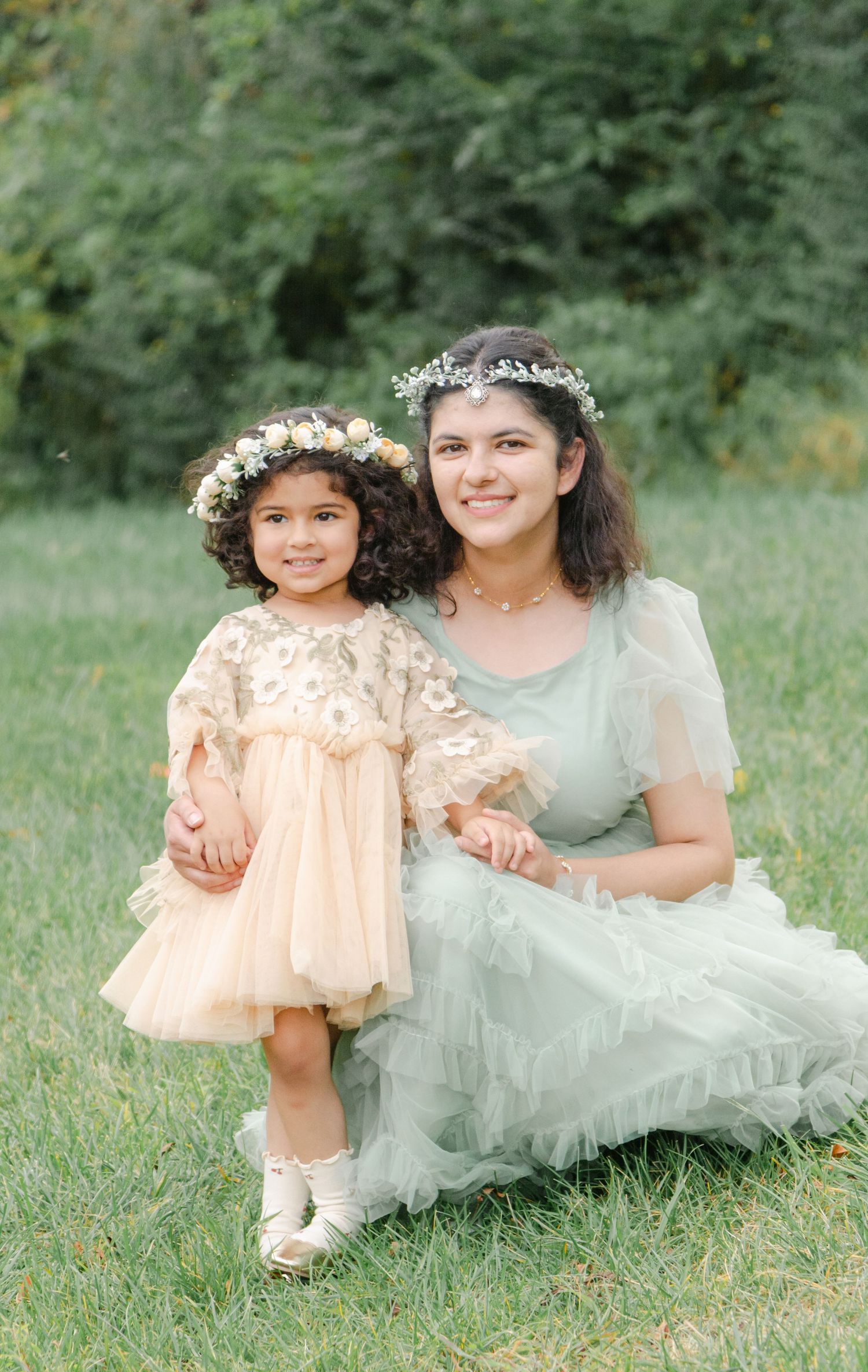 Two people in soft pastel dresses sit together on grass during an outdoor photo session.