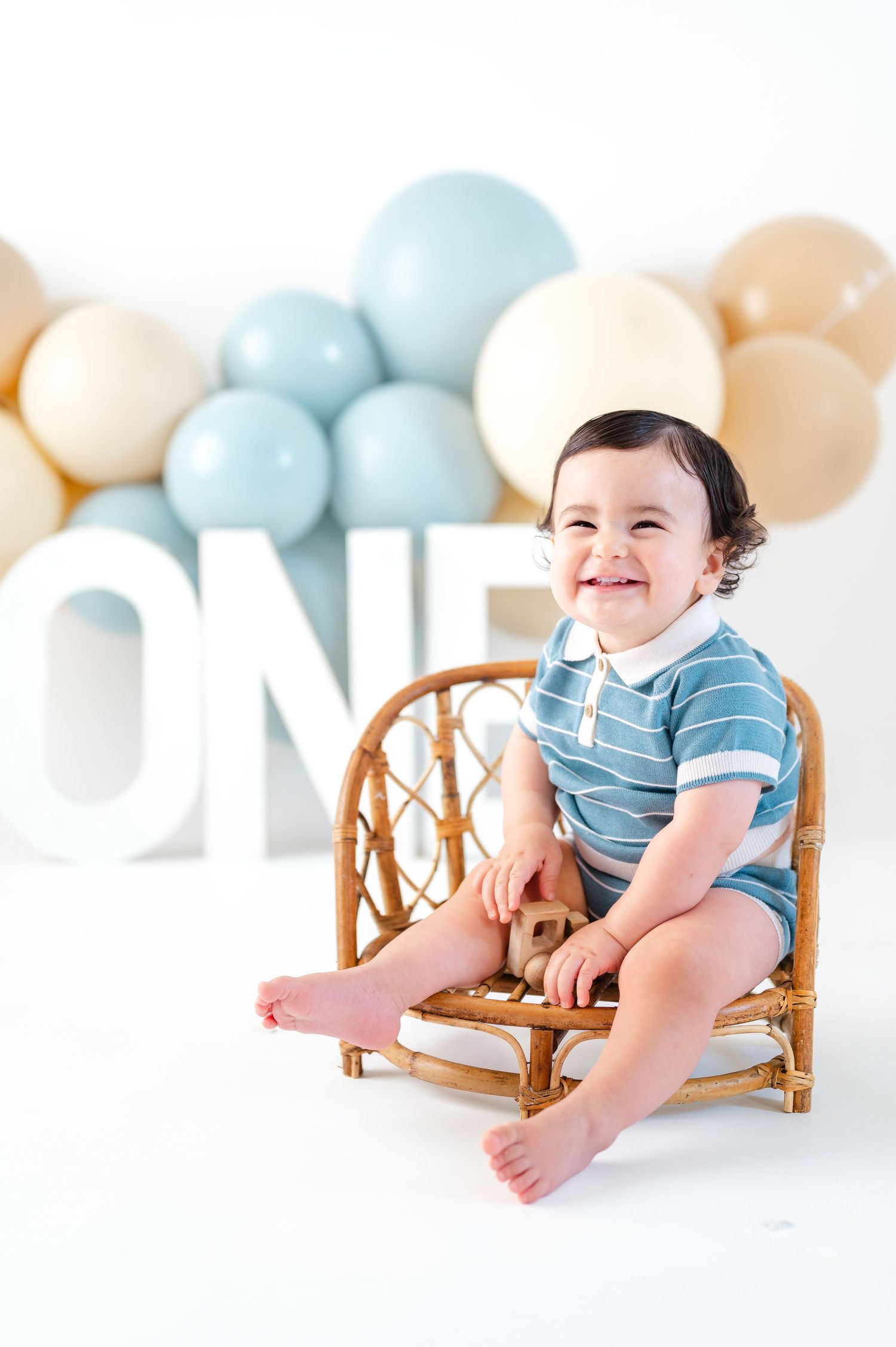 A smiling baby in a blue romper sits in a rattan chair with ONE letters and pastel balloons for a first birthday.