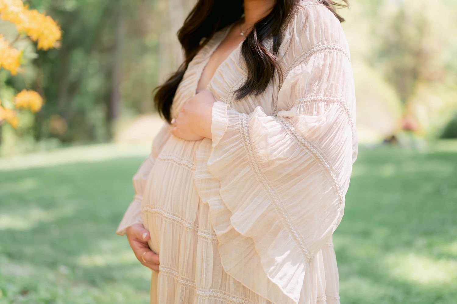 Pregnant woman in flowing white dress cradling baby bump outdoors in lush green garden setting.