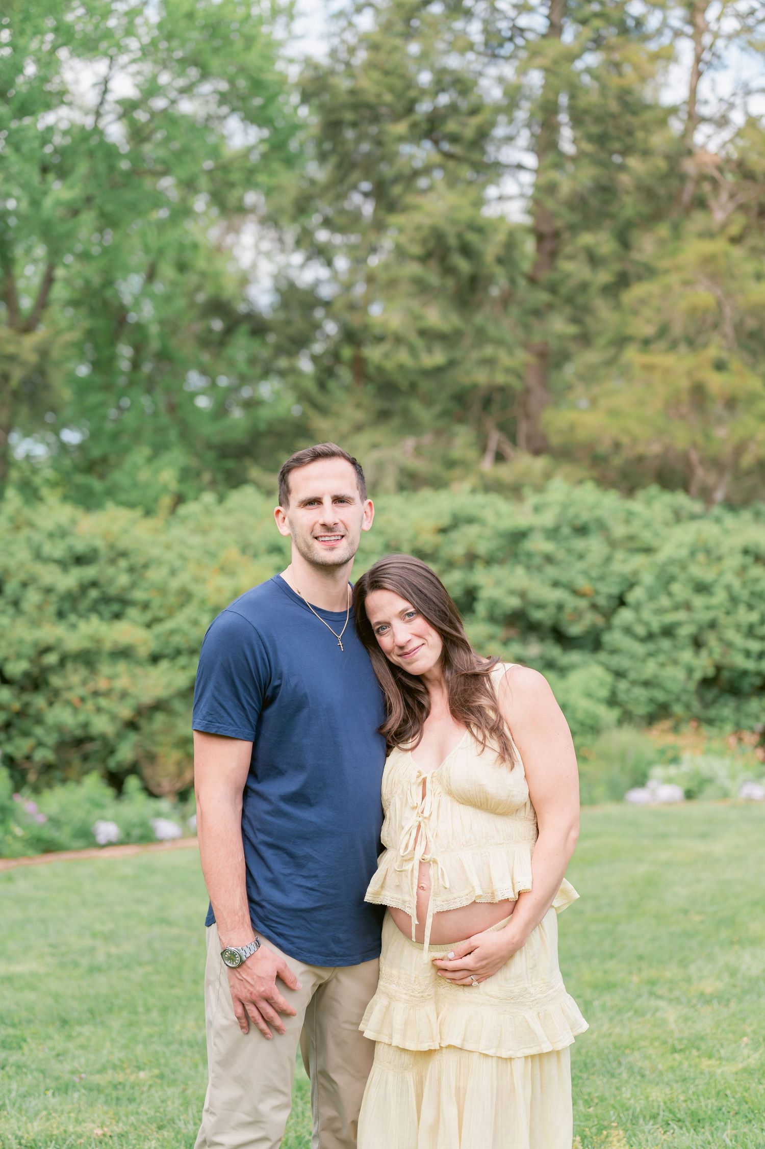 A couple poses outdoors in a garden, the woman in a yellow dress cradling her baby bump during a maternity photo session.