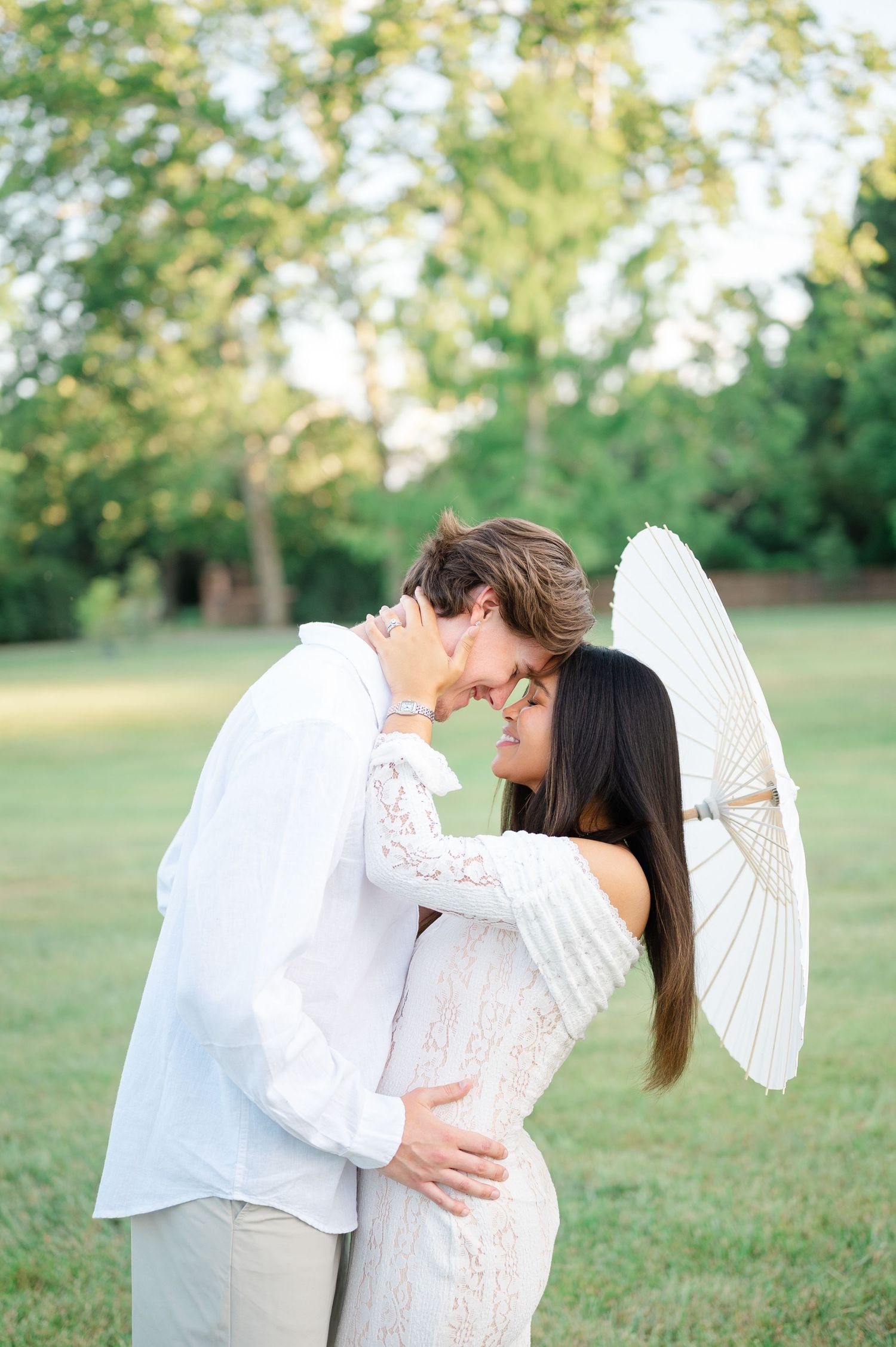 Couple in white outfits sharing a romantic moment outdoors, woman holding a paper parasol in a lush green park.
