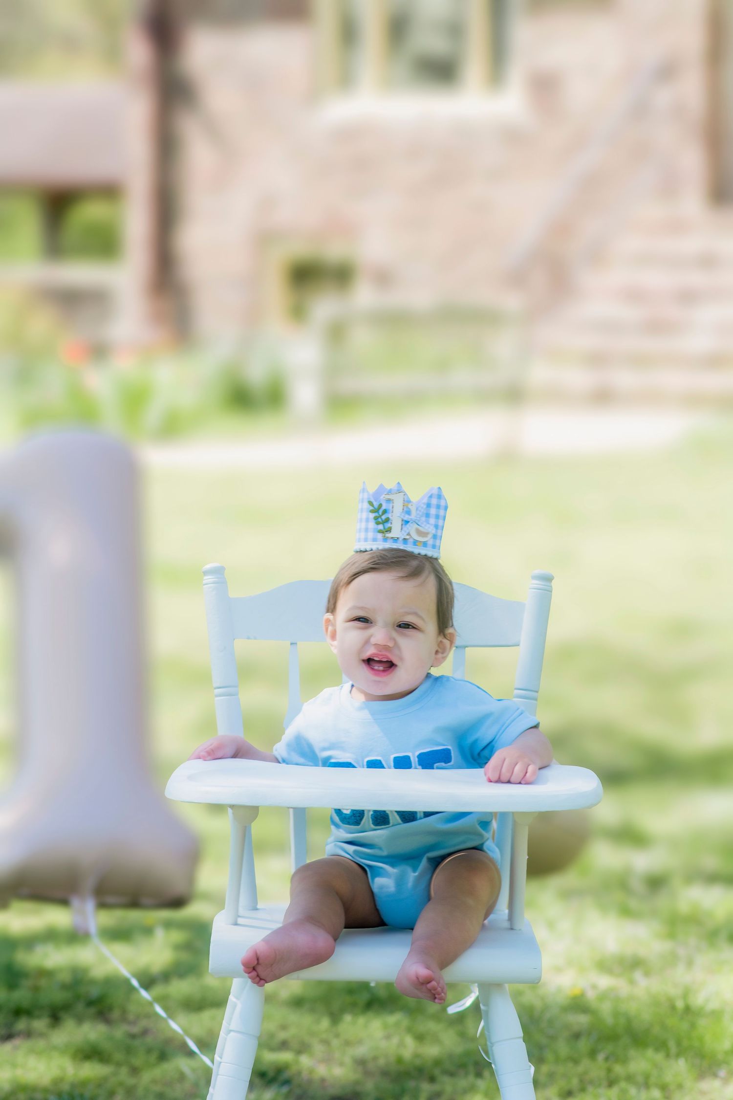 Baby smiles while sitting in antique white high chair on grass during outdoor photoshoot.