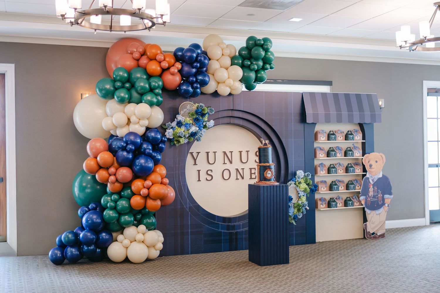 Navy blue and orange balloon arch decorations adorn a modern event space with 'is one' text signage on the wall.
