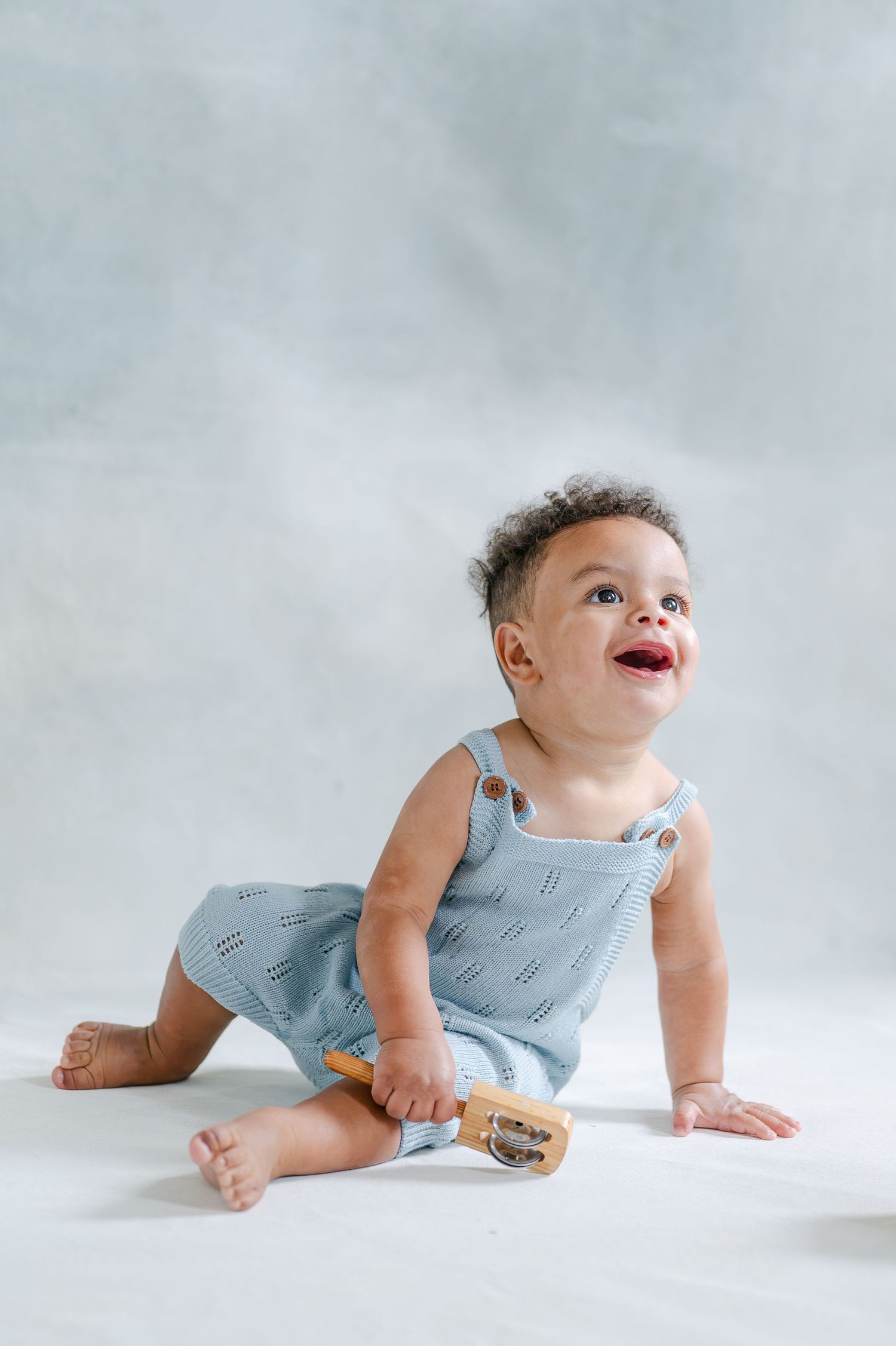 Smiling baby in light blue romper sitting on white background, holding a wooden toy.