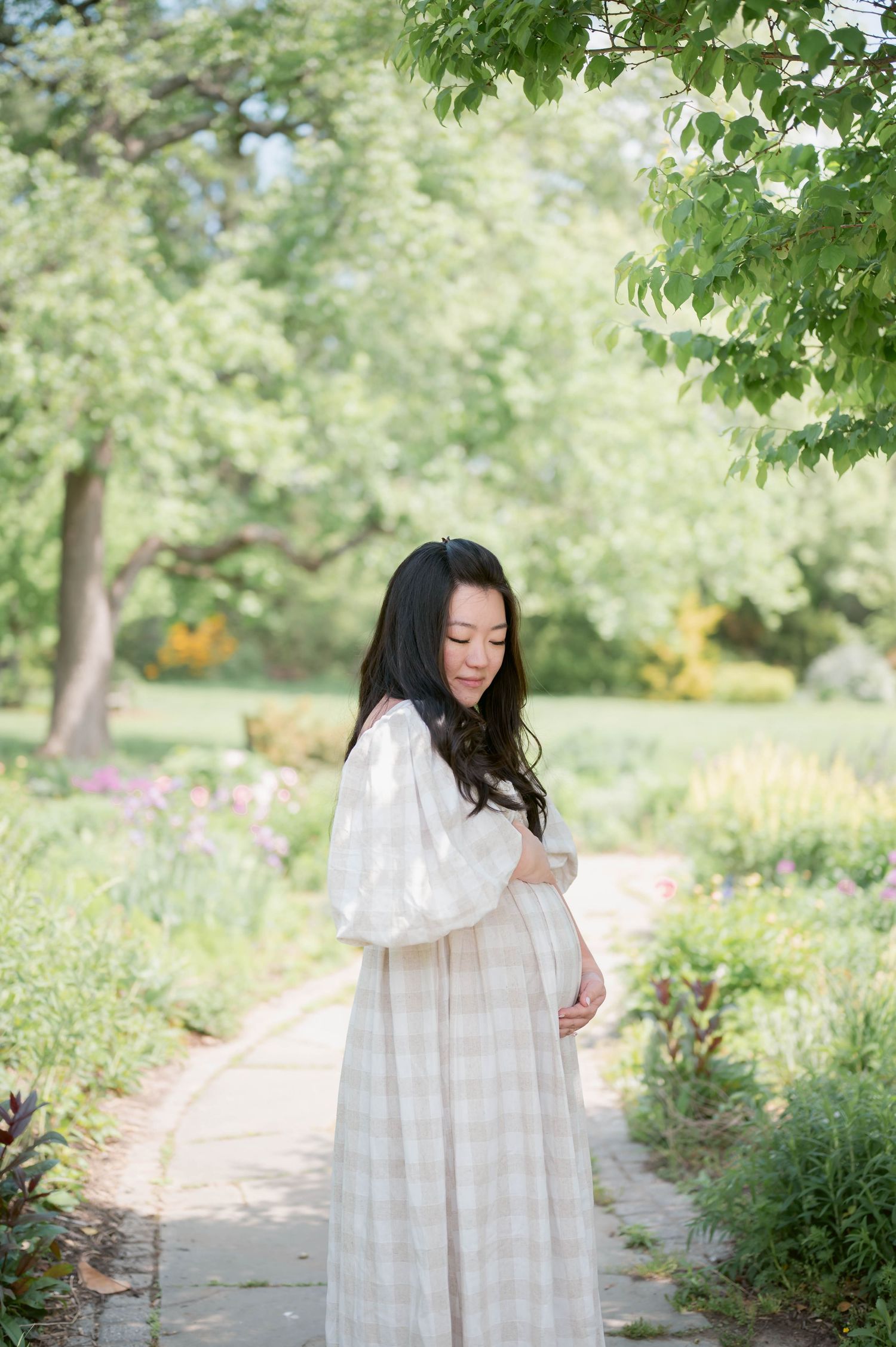 Pregnant woman in white dress holding baby bump on garden path surrounded by lush green trees and colorful flowers.