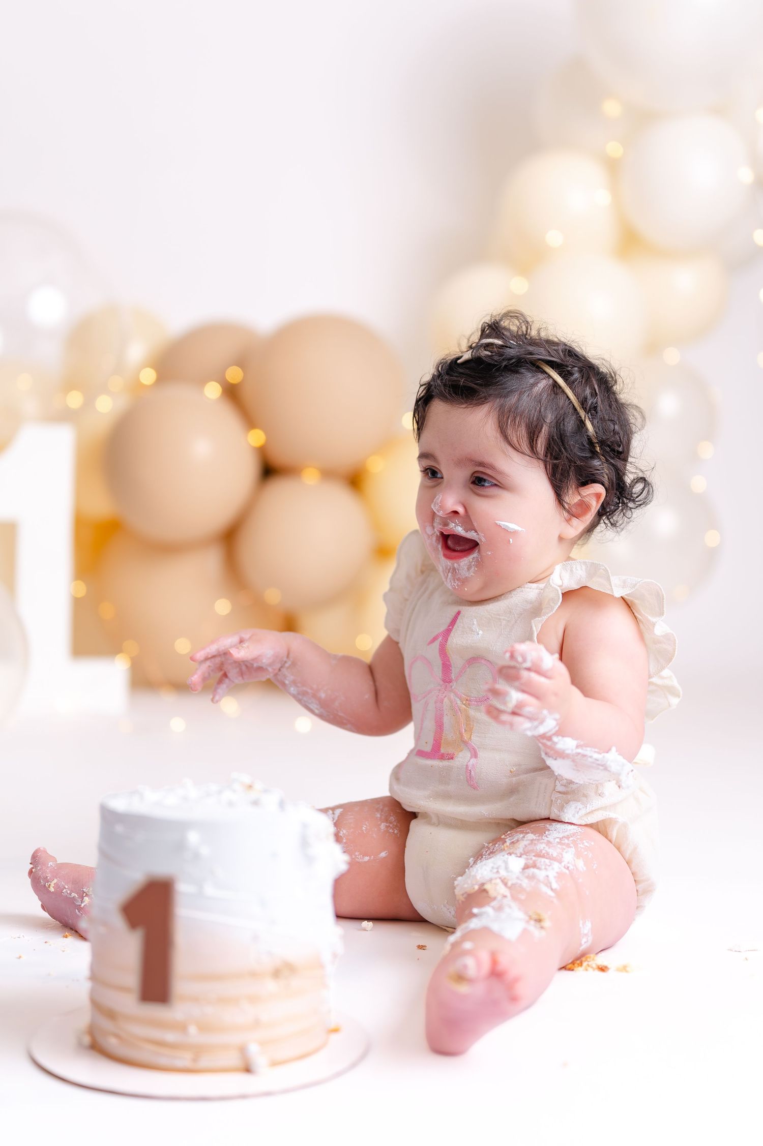 Baby girl smashing her first birthday cake, covered in frosting, surrounded by beige balloons and fairy lights.