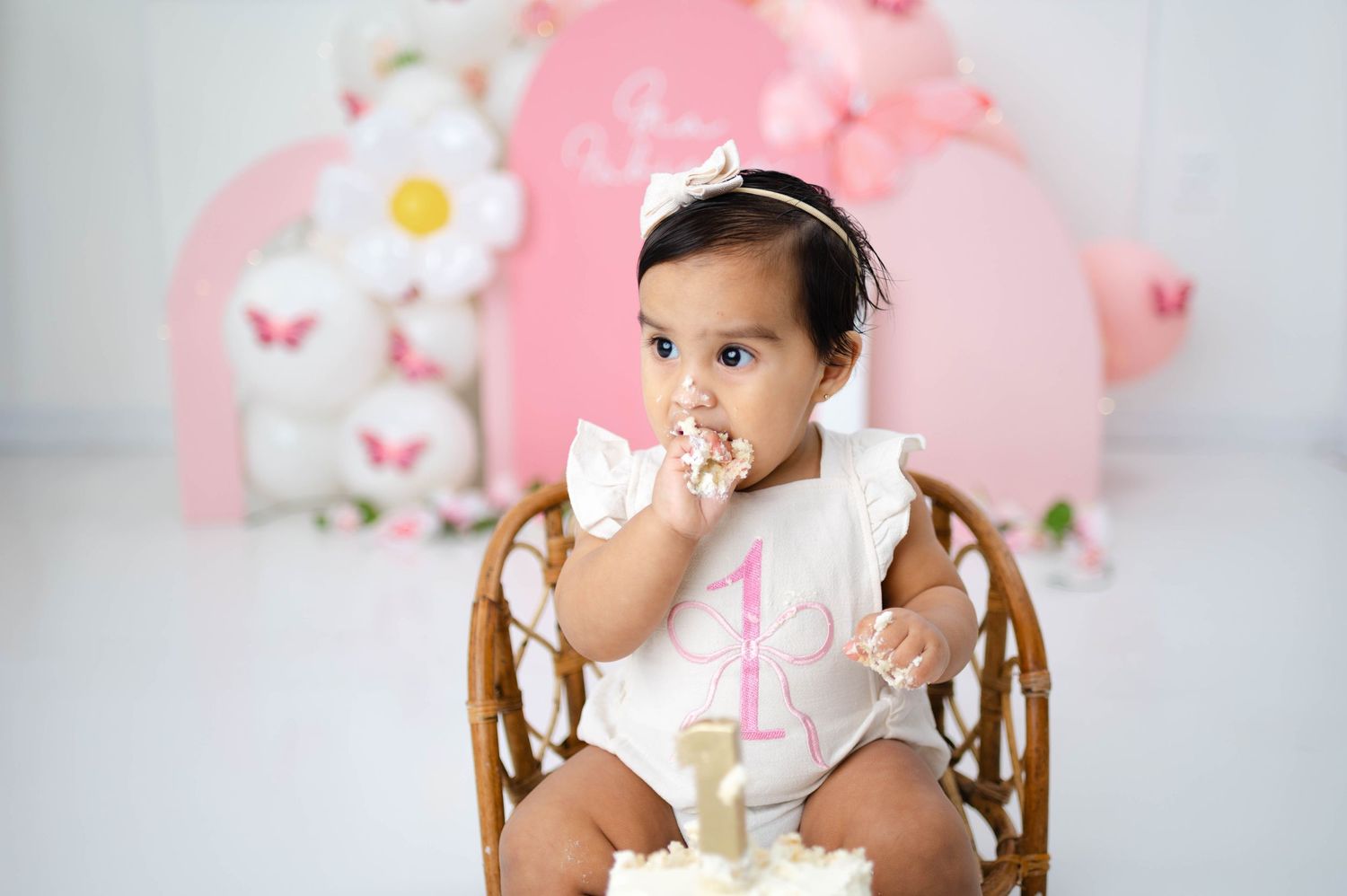 Baby girl in white dress tastes birthday cake while seated in vintage wooden high chair against pink backdrop.