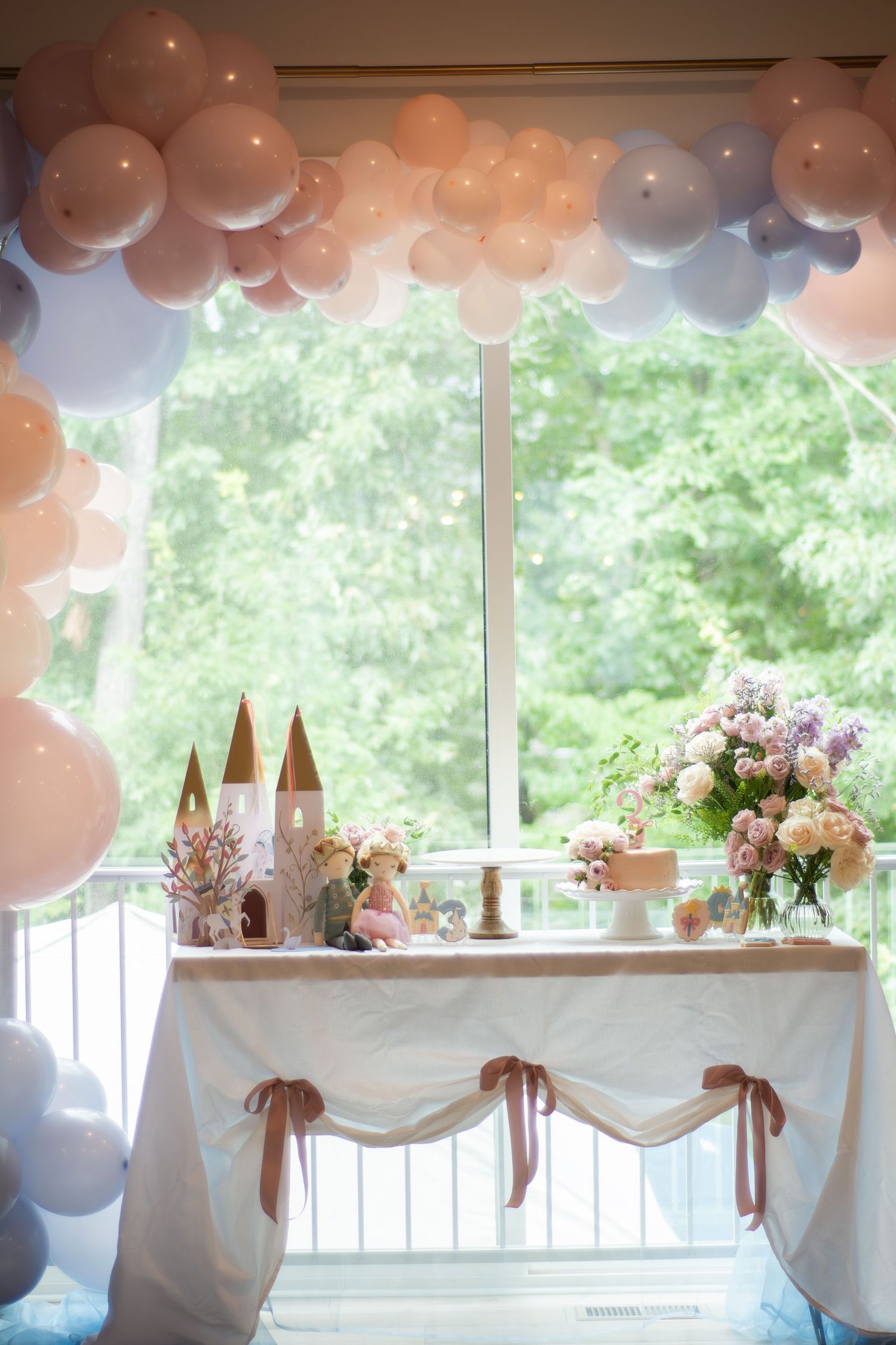 Elegant party setup with white draped tables, rose gold balloons, and floral centerpieces in a bright windowed venue.