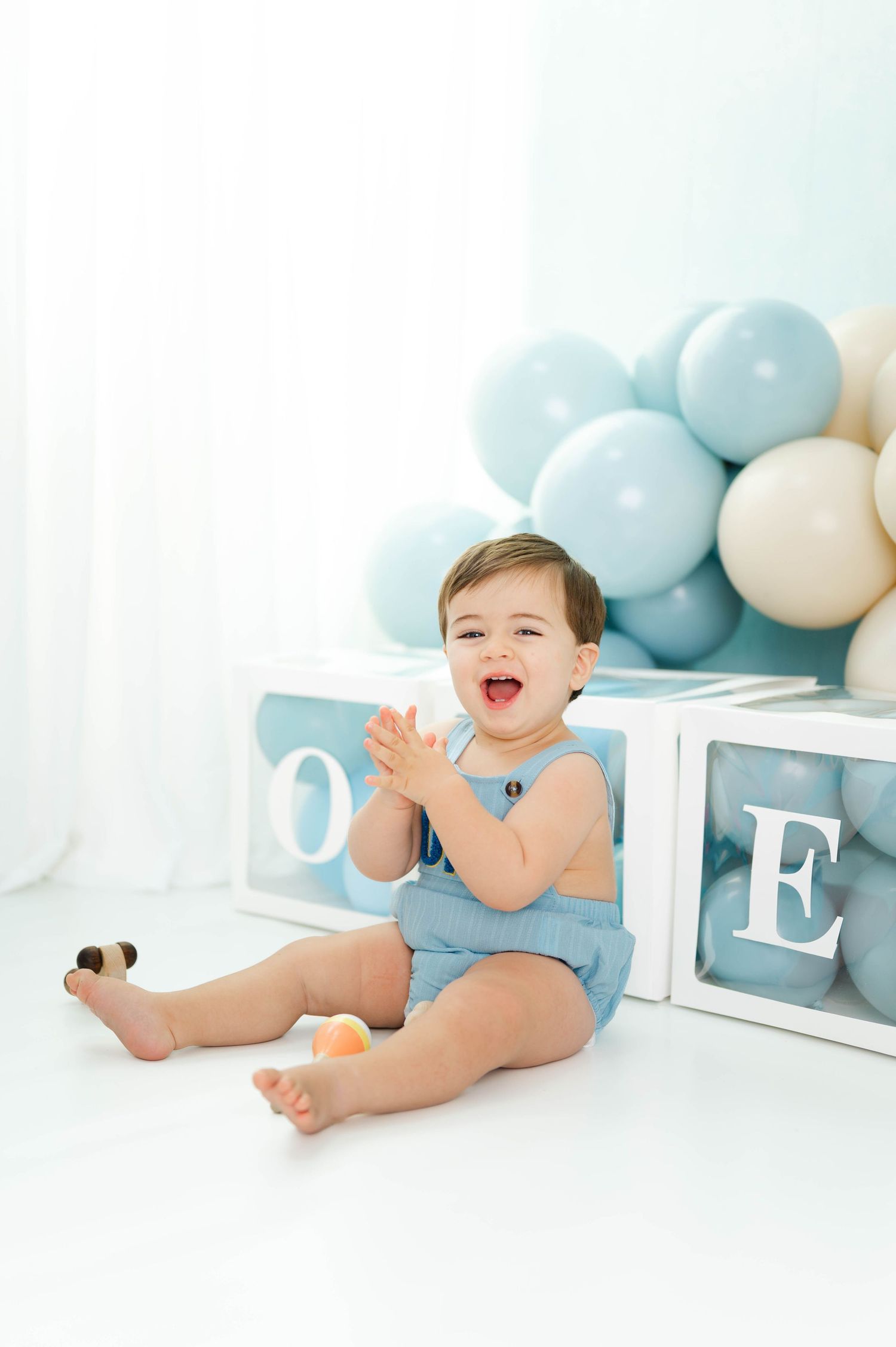 A baby in blue romper sits on white floor with decorative blocks and pastel blue balloons in the background.