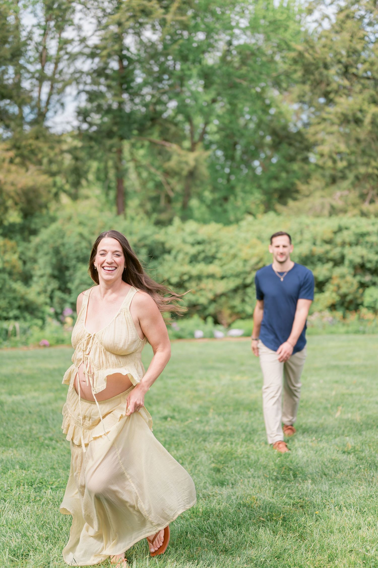 A smiling woman in a yellow dress runs through a lush green park as a man in a navy shirt follows behind her.