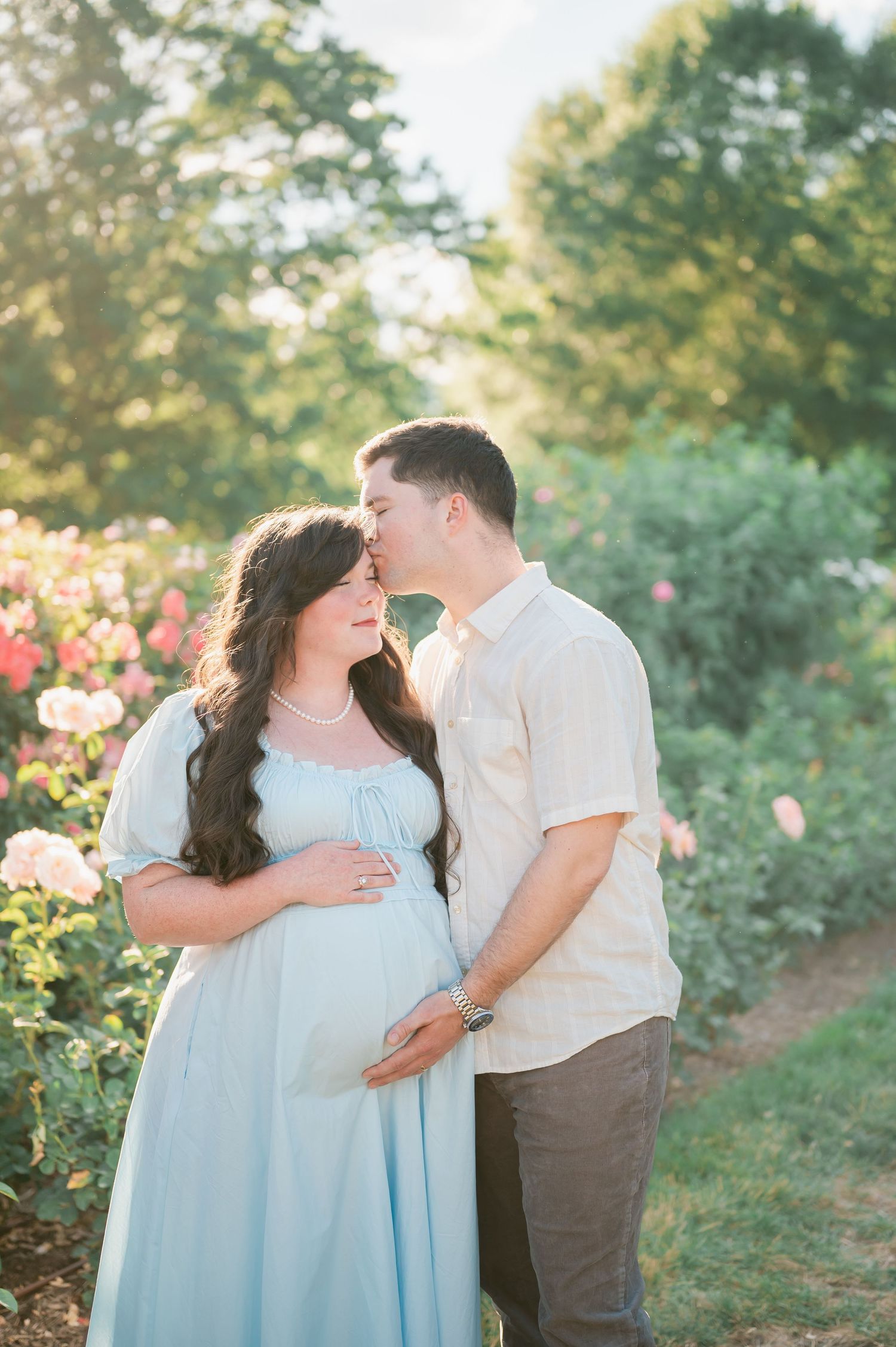 Pregnant woman in light blue dress with partner kissing her forehead in a sunlit rose garden.