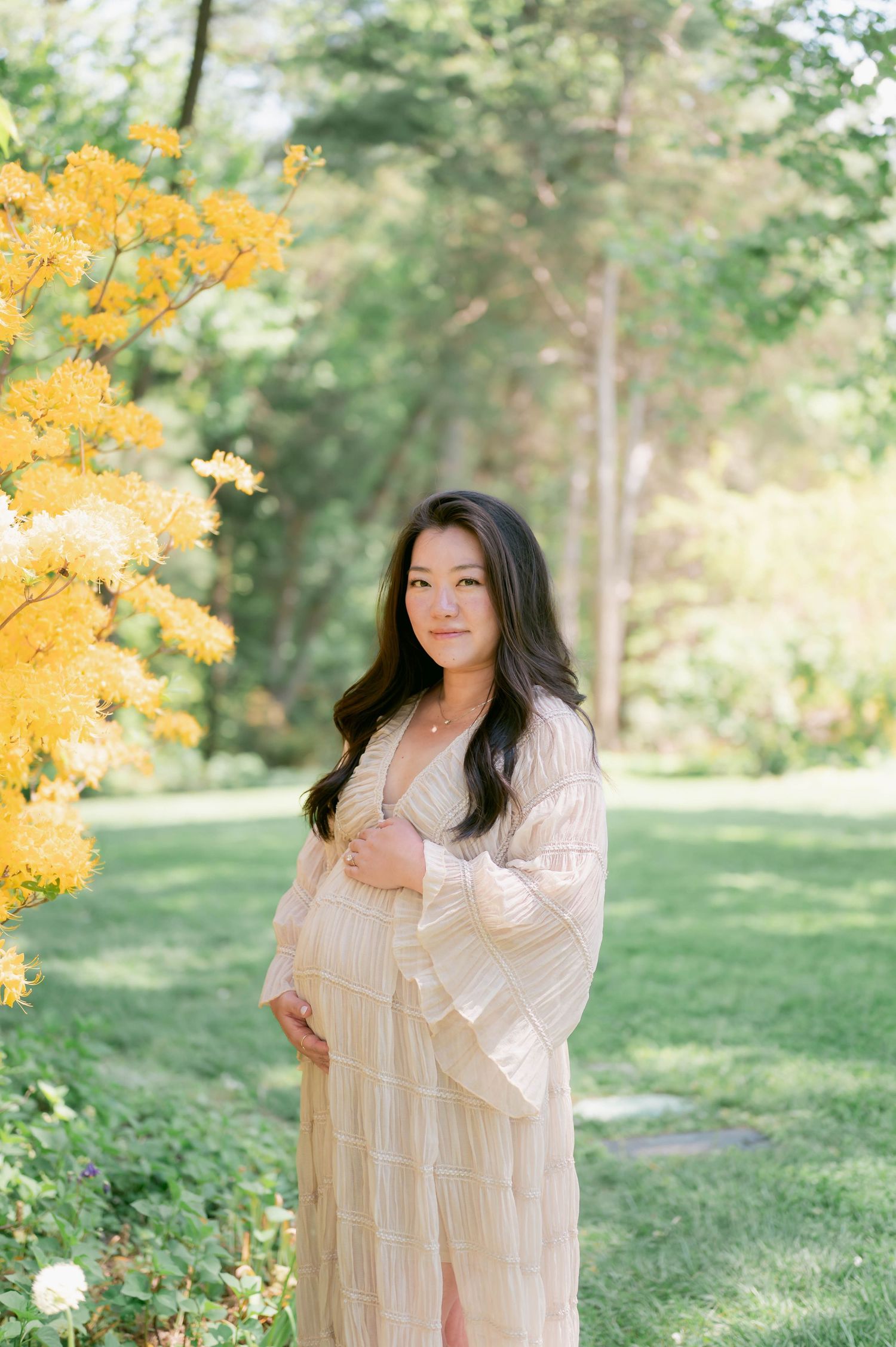 Pregnant woman in cream dress poses near yellow blooming tree in lush green garden during maternity photo session.
