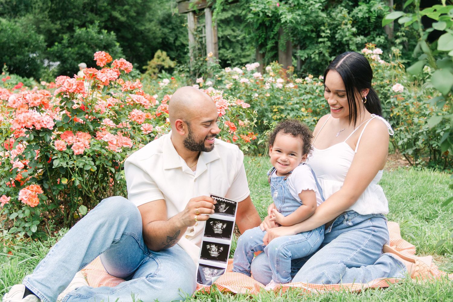 Family lying on grass in a rose garden sharing special moments with ultrasound photos.