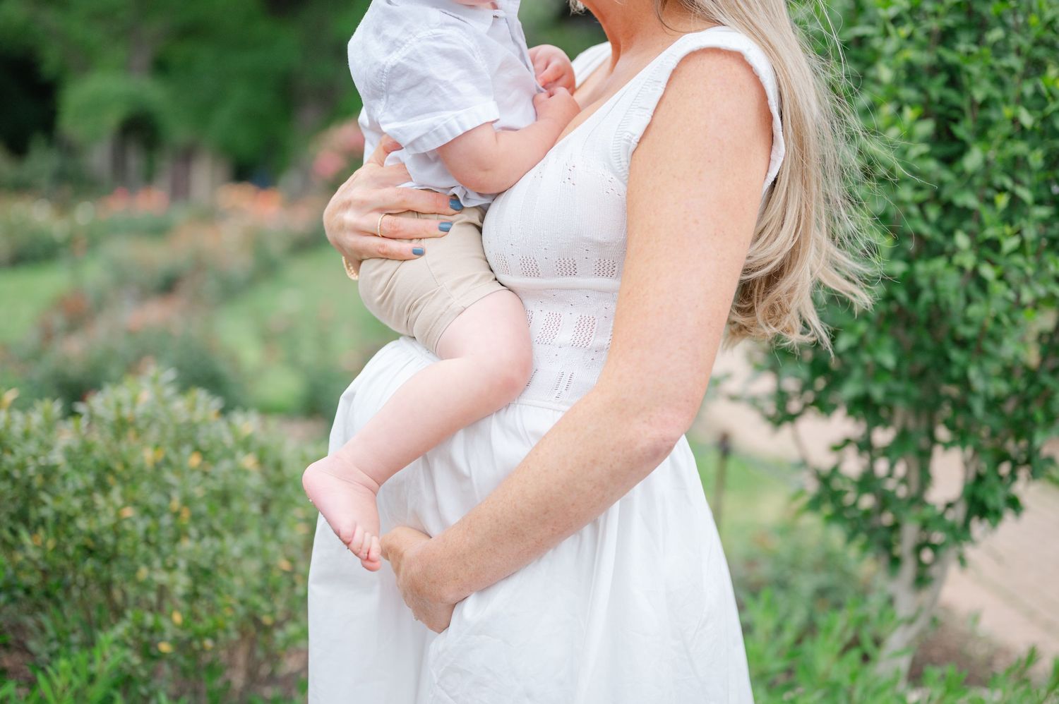 Pregnant woman in white dress holding baby outdoors in garden setting.