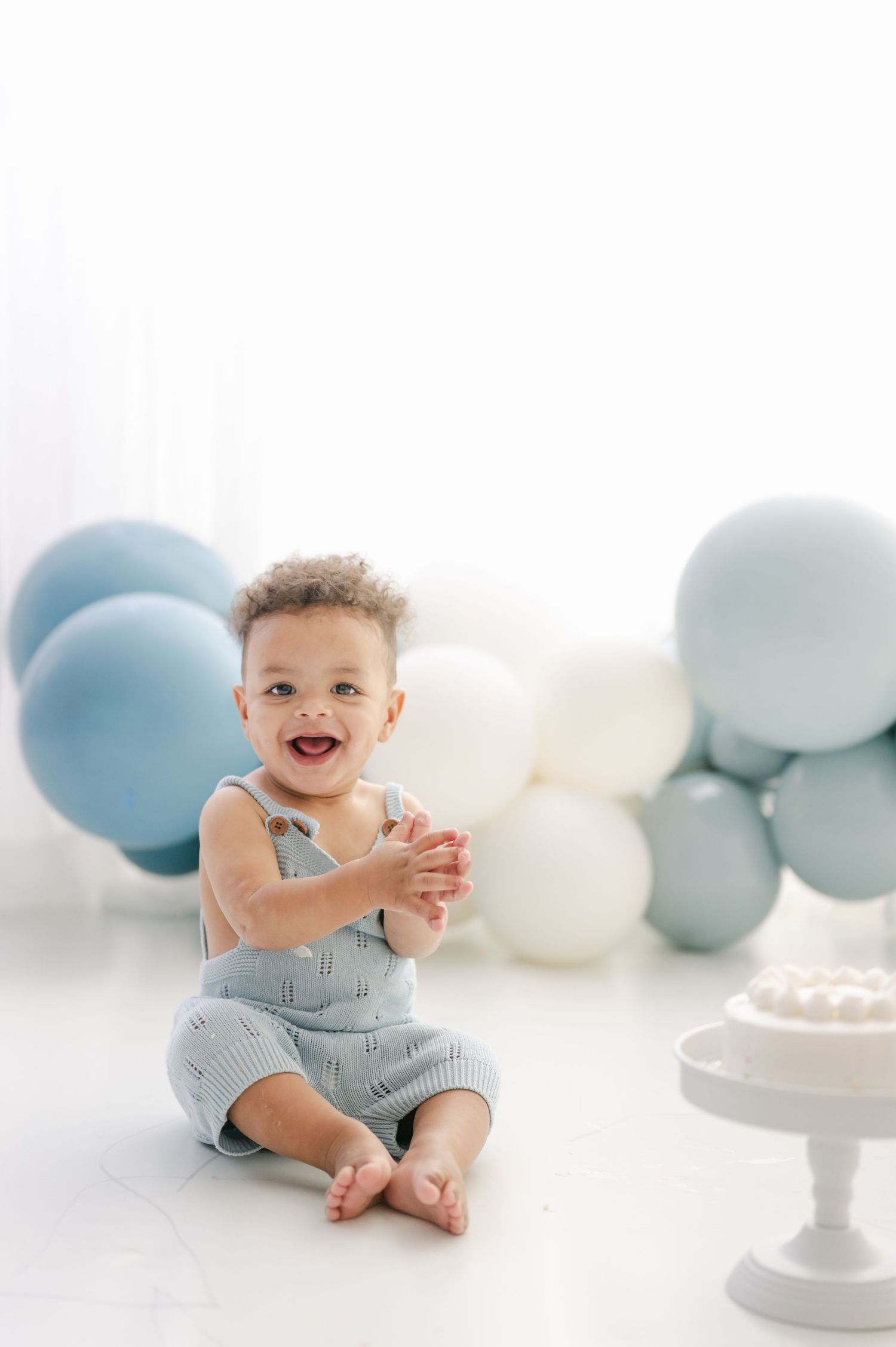 Happy baby boy in blue romper clapping hands at first birthday cake smash with blue and white balloons.
