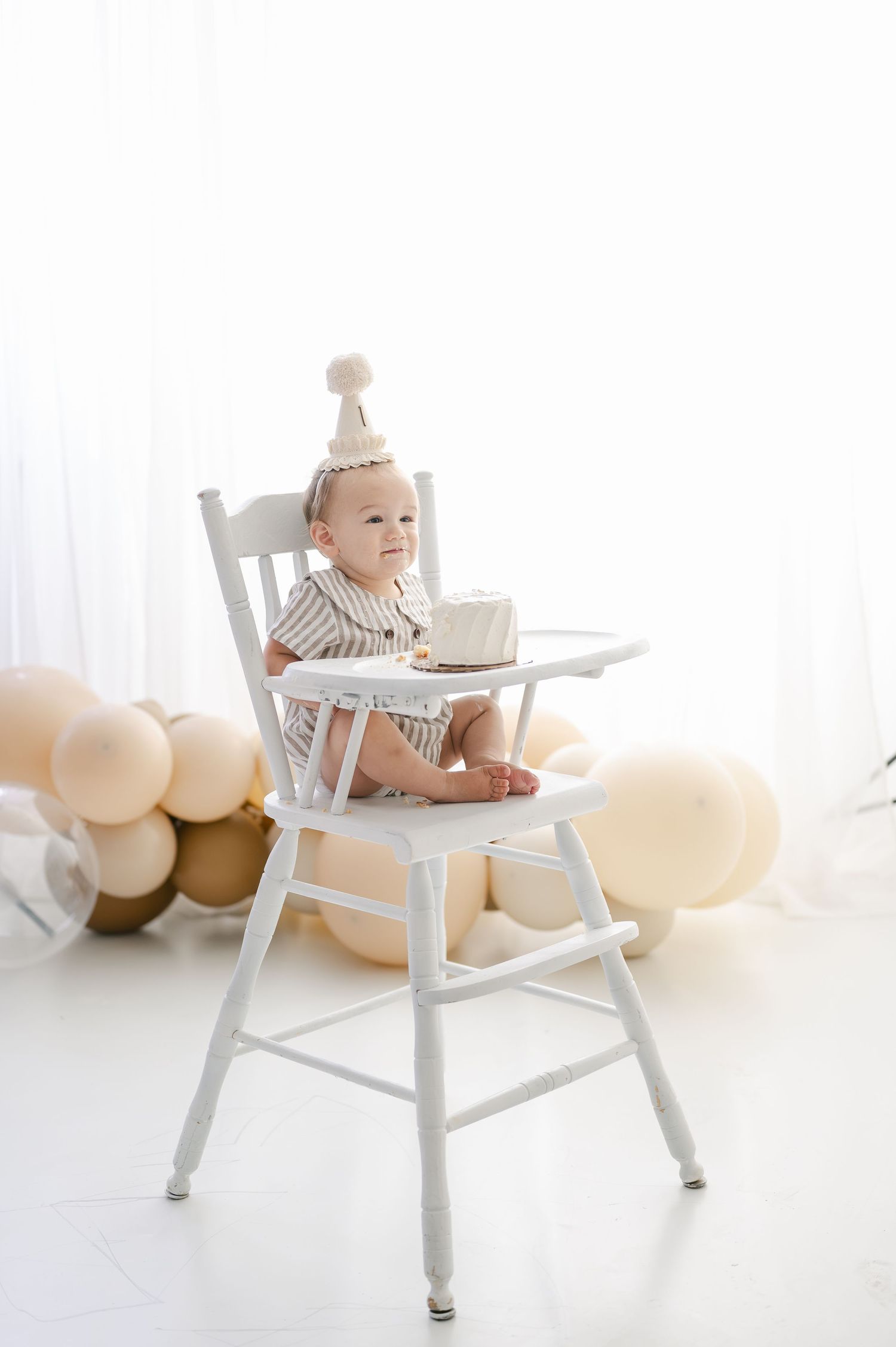 A baby in a striped outfit and party hat sits in a white high chair with a small cake, surrounded by neutral balloons.