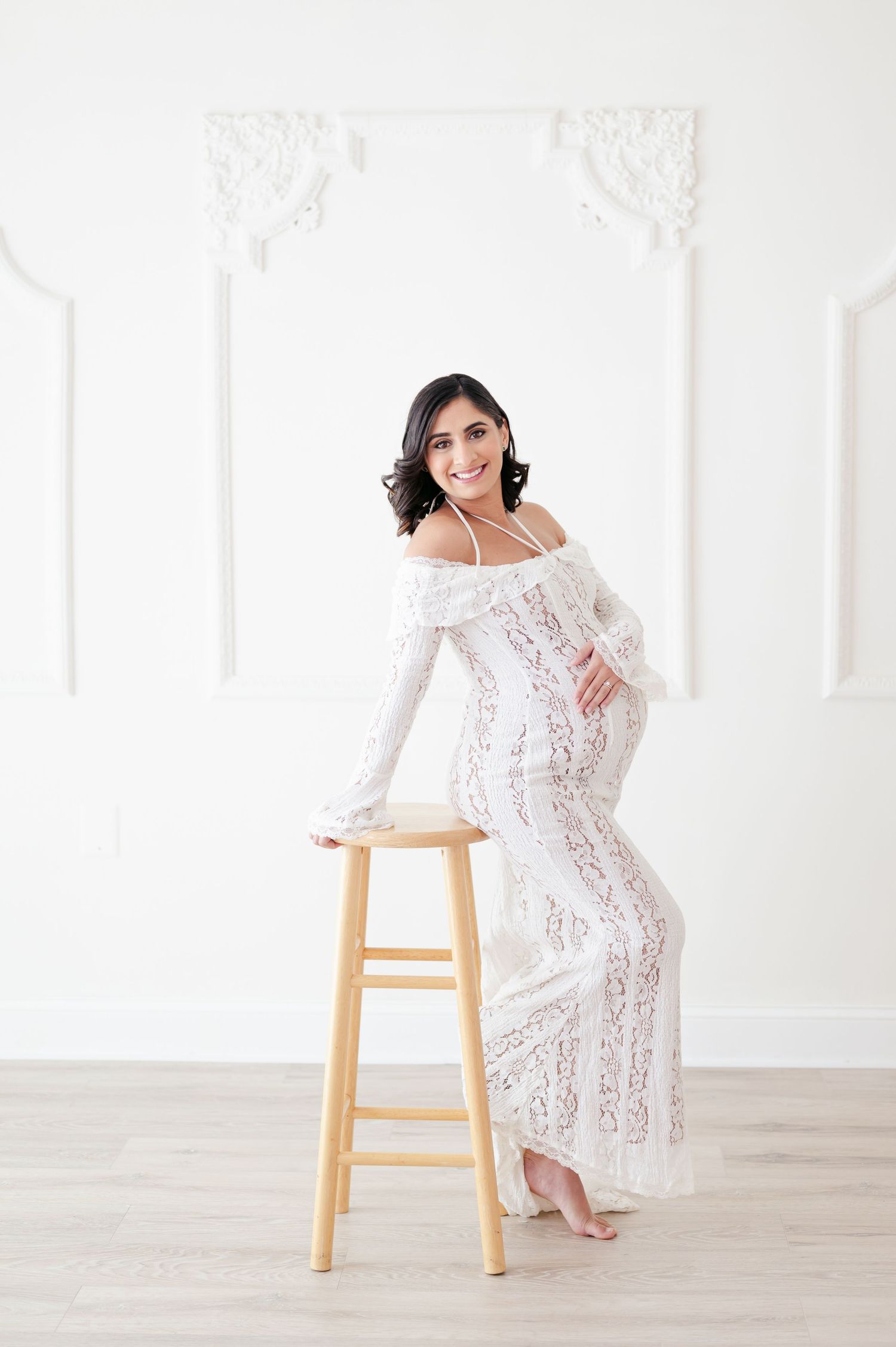 A smiling woman in a white lace off-shoulder gown leans gracefully on a wooden stool in a bright white studio.