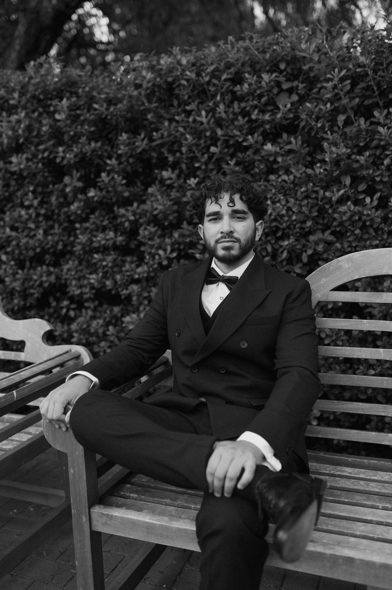 Black and white portrait of person in formal attire seated on wooden bench in garden setting.