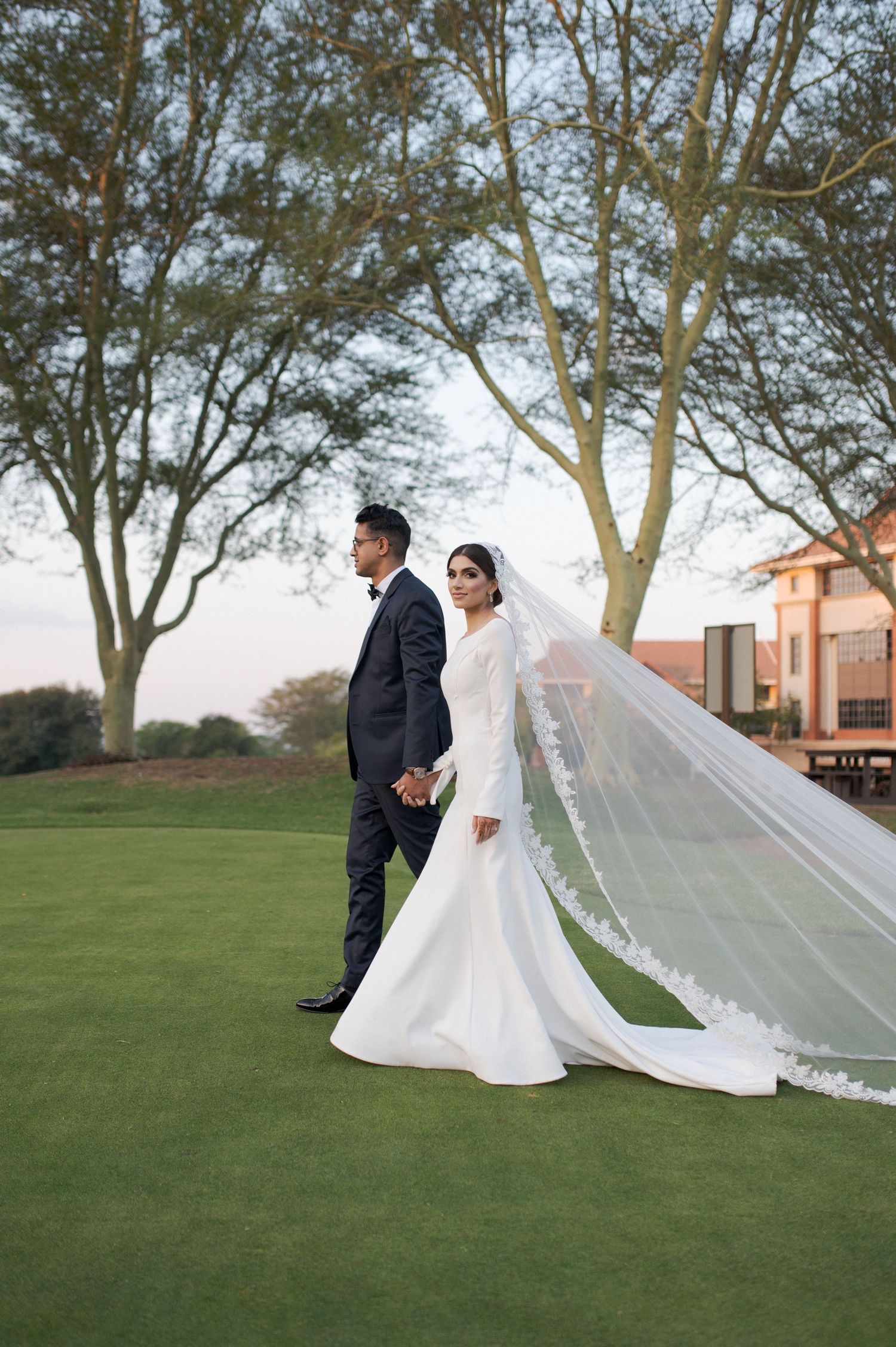 Elegant wedding couple standing on manicured lawn with long flowing veil and modern architecture.