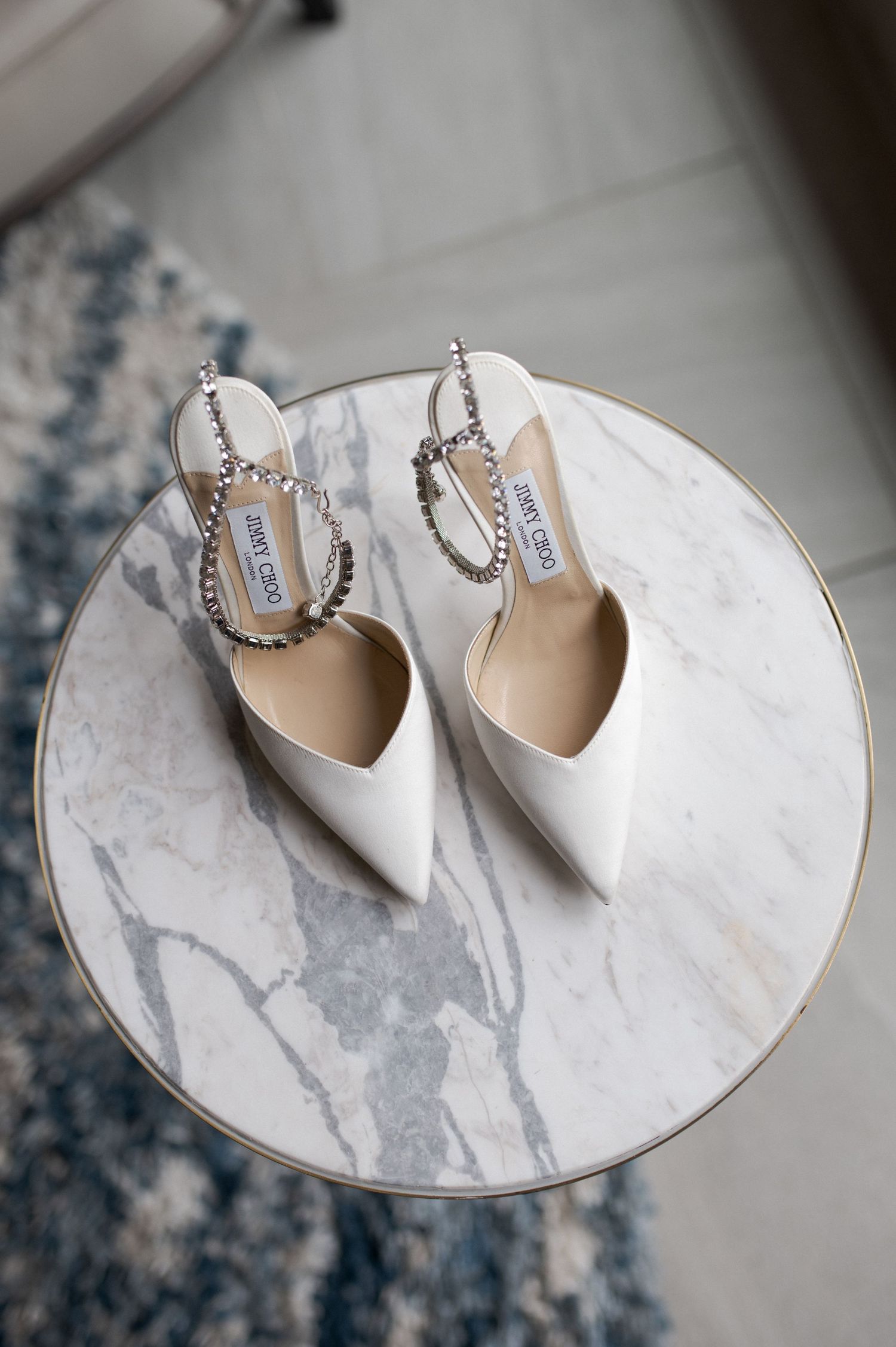 White pointed-toe bridal shoes with crystal straps displayed on a marble table.