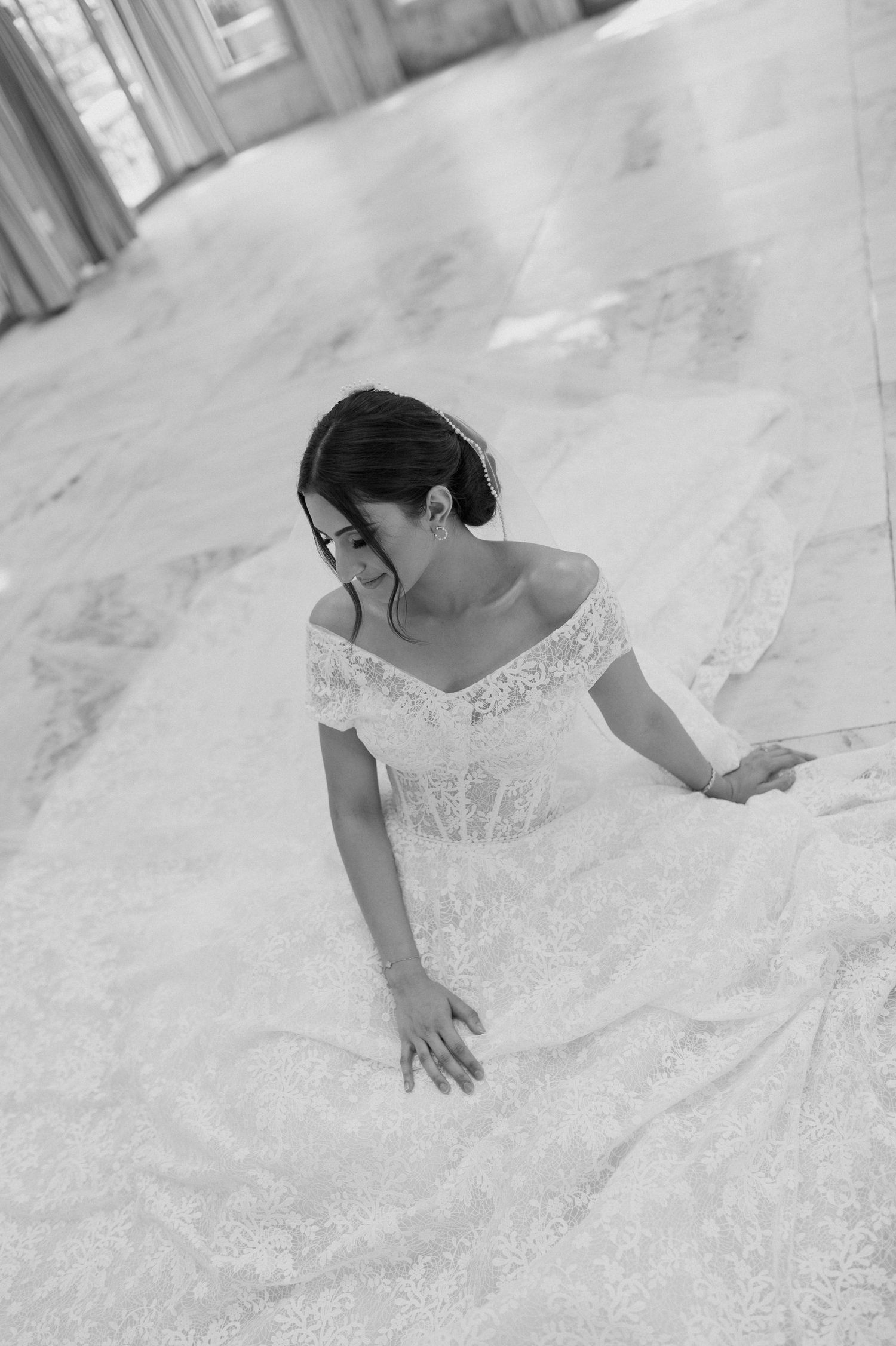 Black and white portrait of bride in delicate lace gown sitting gracefully on marble floor.